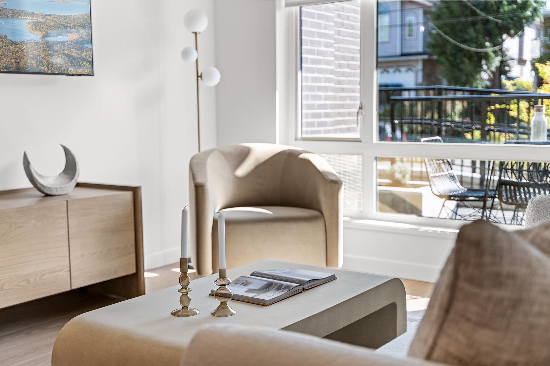 Hawthorne – A sunlit, modern living room corner featuring a plush beige armchair, a minimalist coffee table with tapered candles and an open book, and a low light-wood console cabinet. Large floor-to-ceiling windows let in bright daylight and reveal an adjacent private balcony with outdoor seating.