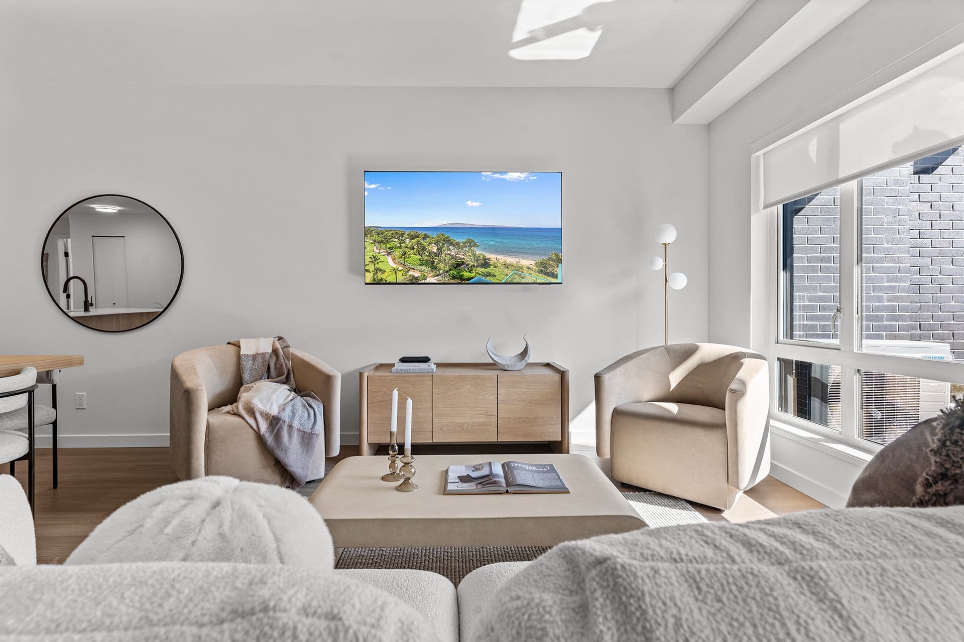 Hawthorne – A modern, bright living room features neutral-toned rounded armchairs, a wooden media console under a wall-mounted television, and a large circular mirror on a plain white wall. Natural light streams in through a large floor-to-ceiling window, highlighting light hardwood floors and plush, textured furniture.