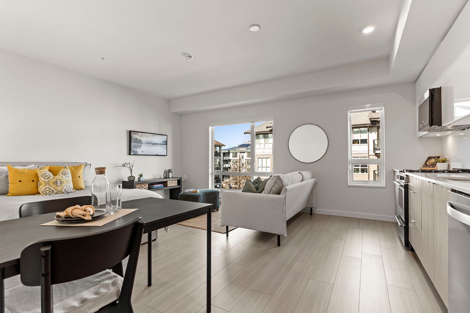 Hawthorne – A bright, open-concept studio apartment featuring light-colored laminate flooring, white walls, and a modern kitchen on the right with light wood lower cabinets and white uppers. The living space includes a light grey sofa facing large windows overlooking neighboring residential buildings, adjacent to a bed styled with vibrant yellow throw pillows.