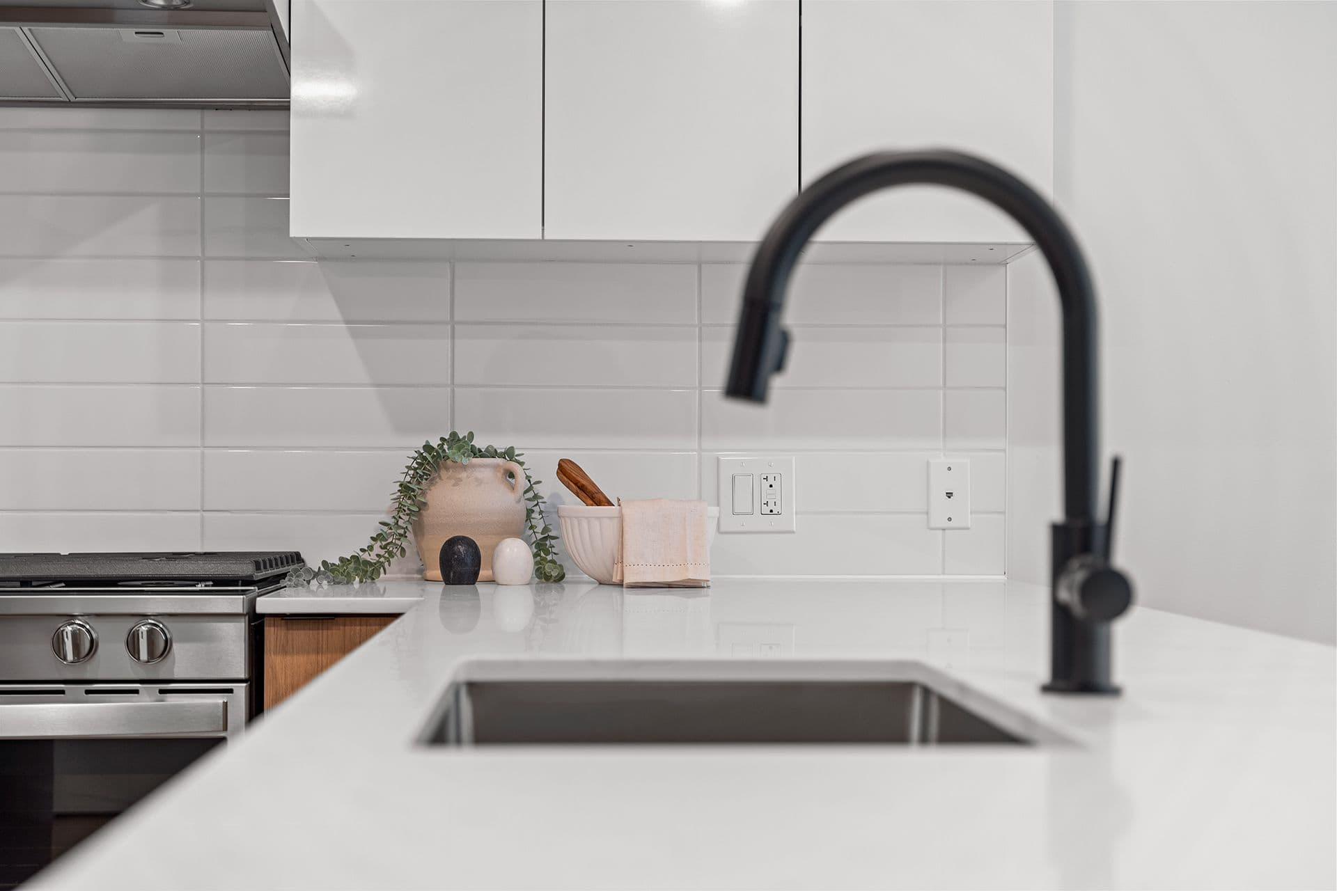 Hawthorne – A close-up view of a modern kitchen counter featuring a matte black gooseneck faucet and an undermount sink set into white quartz countertops. White subway tile backsplash, sleek upper cabinetry, stainless steel range stove, and subtle decorative accents create a bright, clean aesthetic.