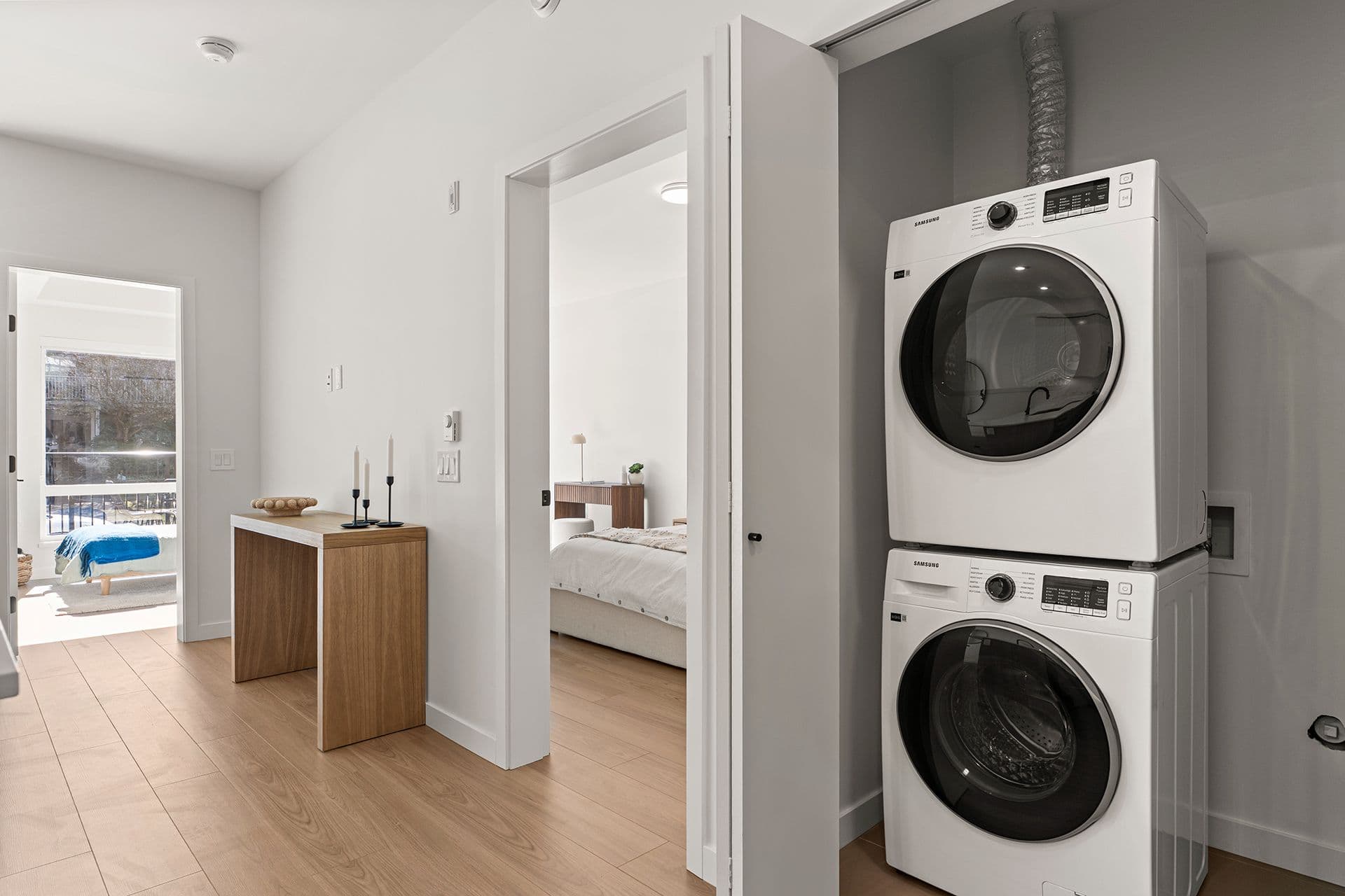 Hawthorne – A modern suite hallway featuring a closet with stacked white Samsung front-loading washer and dryer units next to light-toned hardwood flooring. An open doorway leads into a bright bedroom, while another doorway offers a view toward a sunlit balcony.