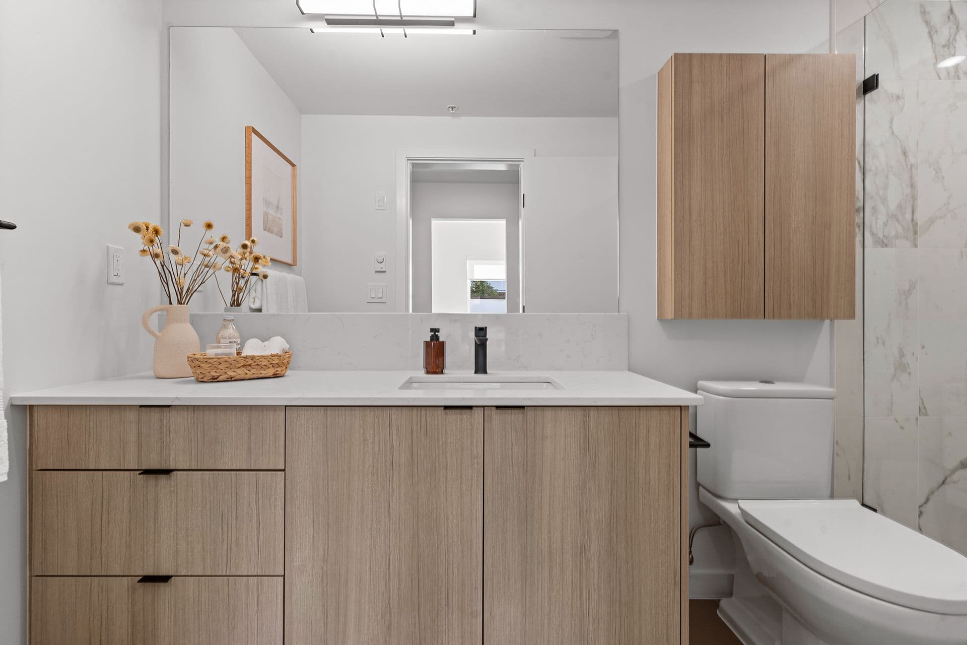 Hawthorne – A modern bathroom featuring a light wood vanity with a white quartz countertop, matte black hardware, and a large frameless mirror above. To the right, a toilet and a glass-enclosed shower with marble-look wall tiles complete the clean, contemporary space.