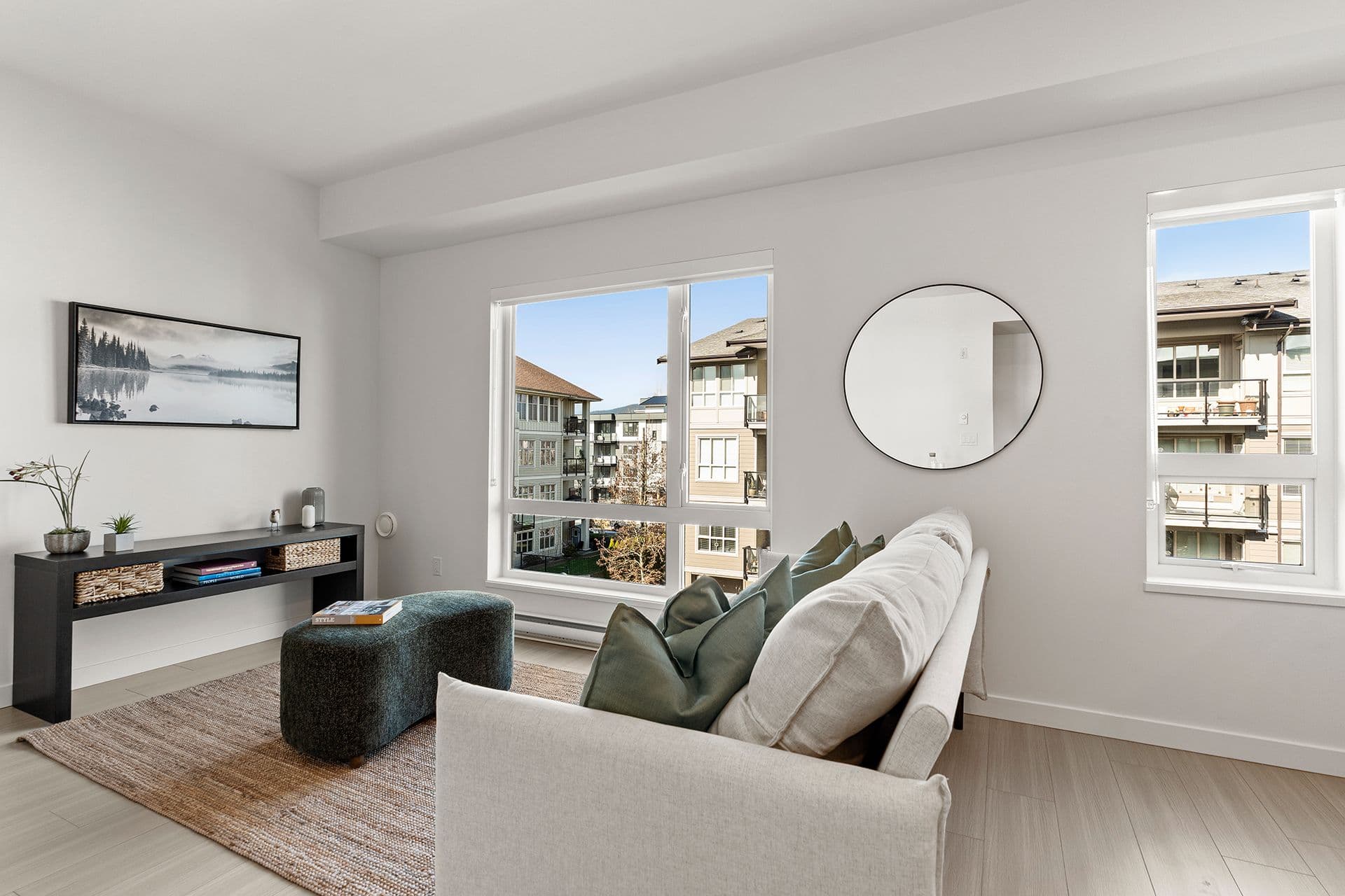 Hawthorne – A modern, bright living room features a white fabric sofa accented with sage green pillows, a low dark wood console table, and a plush dark blue ottoman resting on a woven area rug. Large windows and a large circular wall mirror brighten the white space and offer views of neighboring residential buildings under a clear blue sky.