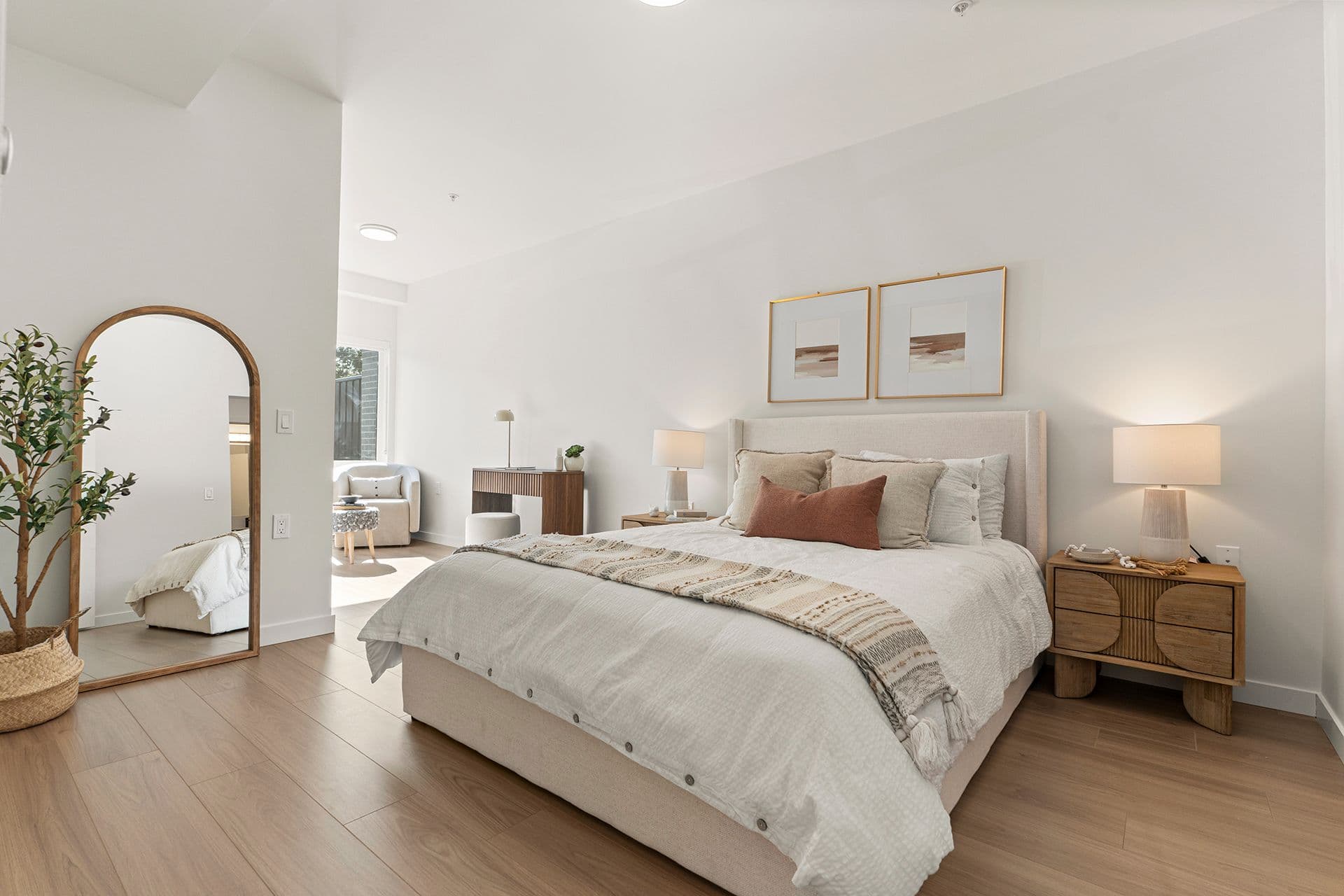 Hawthorne – A bright, modern bedroom styled in neutral tones features a padded upholstered bed with light linens, a terracotta accent pillow, and wooden nightstands with glowing bedside lamps. Natural light streams in from an adjacent sitting nook, illuminating the light oak laminate flooring and a tall arched floor mirror beside a potted plant.
