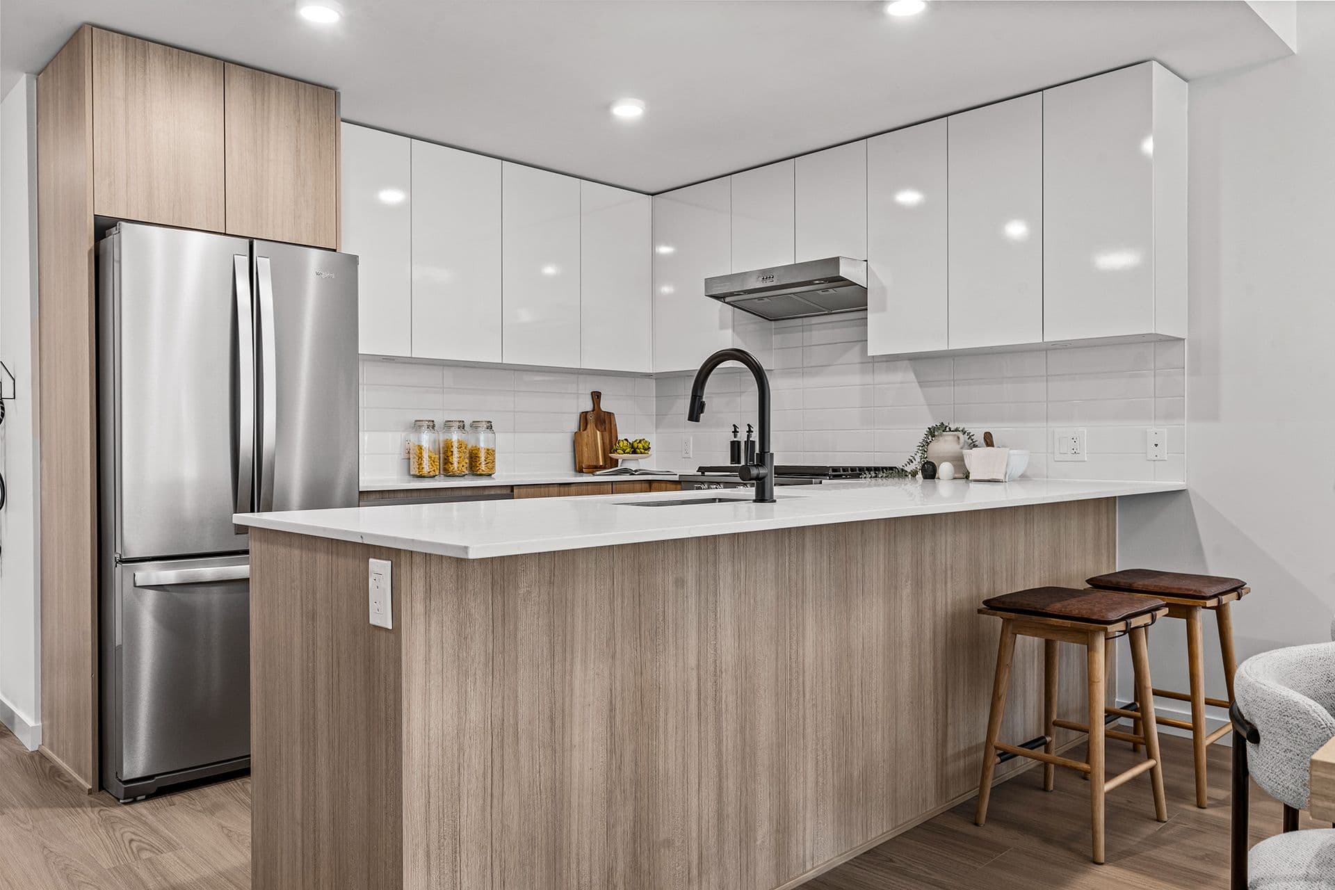 Hawthorne – A modern residential kitchen featuring flat-panel white upper cabinets paired with natural wood grain lower cabinets and island detailing. The space includes bright white quartz countertops, a matte black high-arc faucet, stainless steel appliances, and two leather-topped wooden barstools at the island.