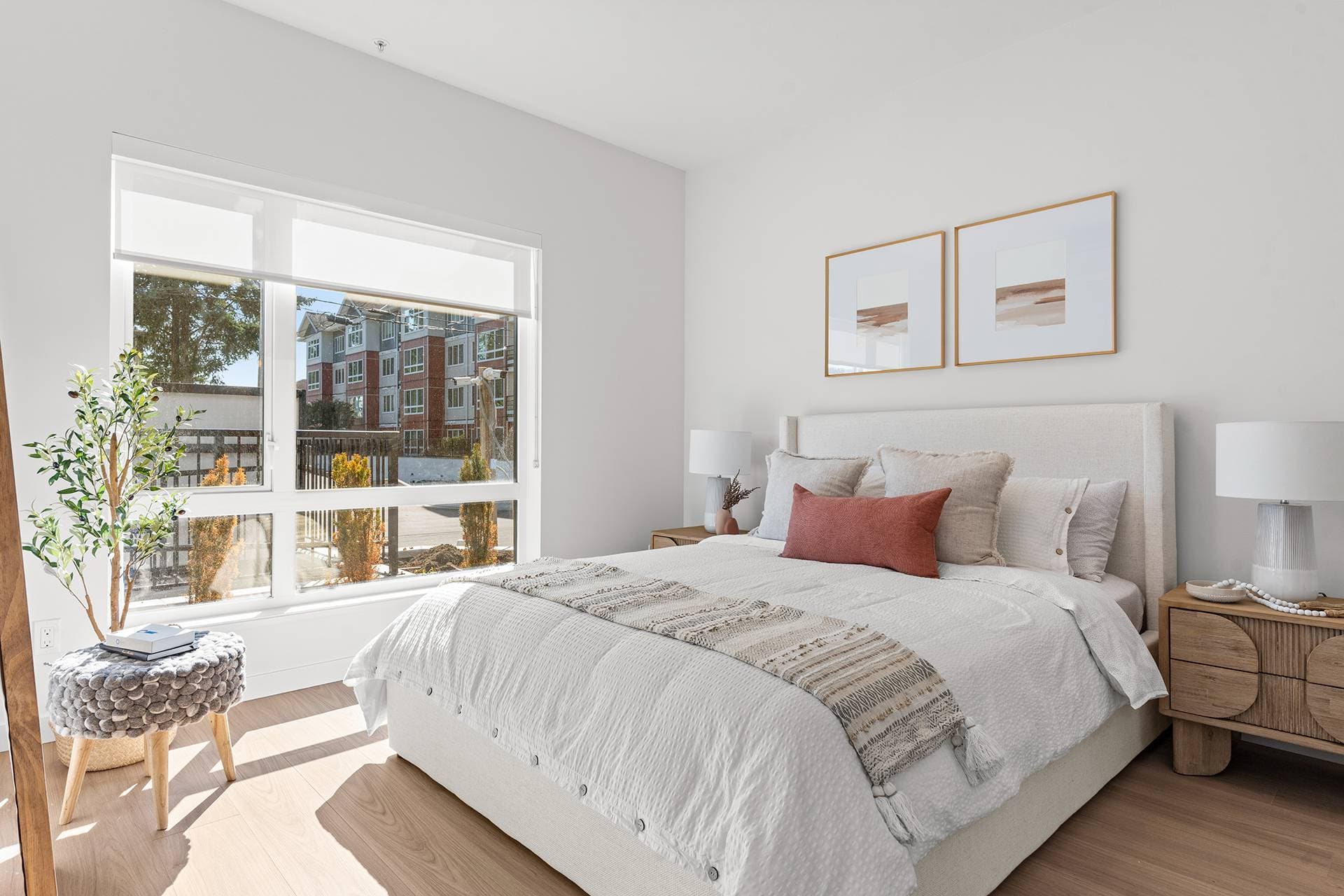 Hawthorne – A modern bedroom filled with bright natural light featuring a neatly dressed bed with neutral linens and a rust-orange accent pillow. Wooden nightstands with matching lamps flank the bed, while a large window overlooks a residential street lined with multi-family housing.
