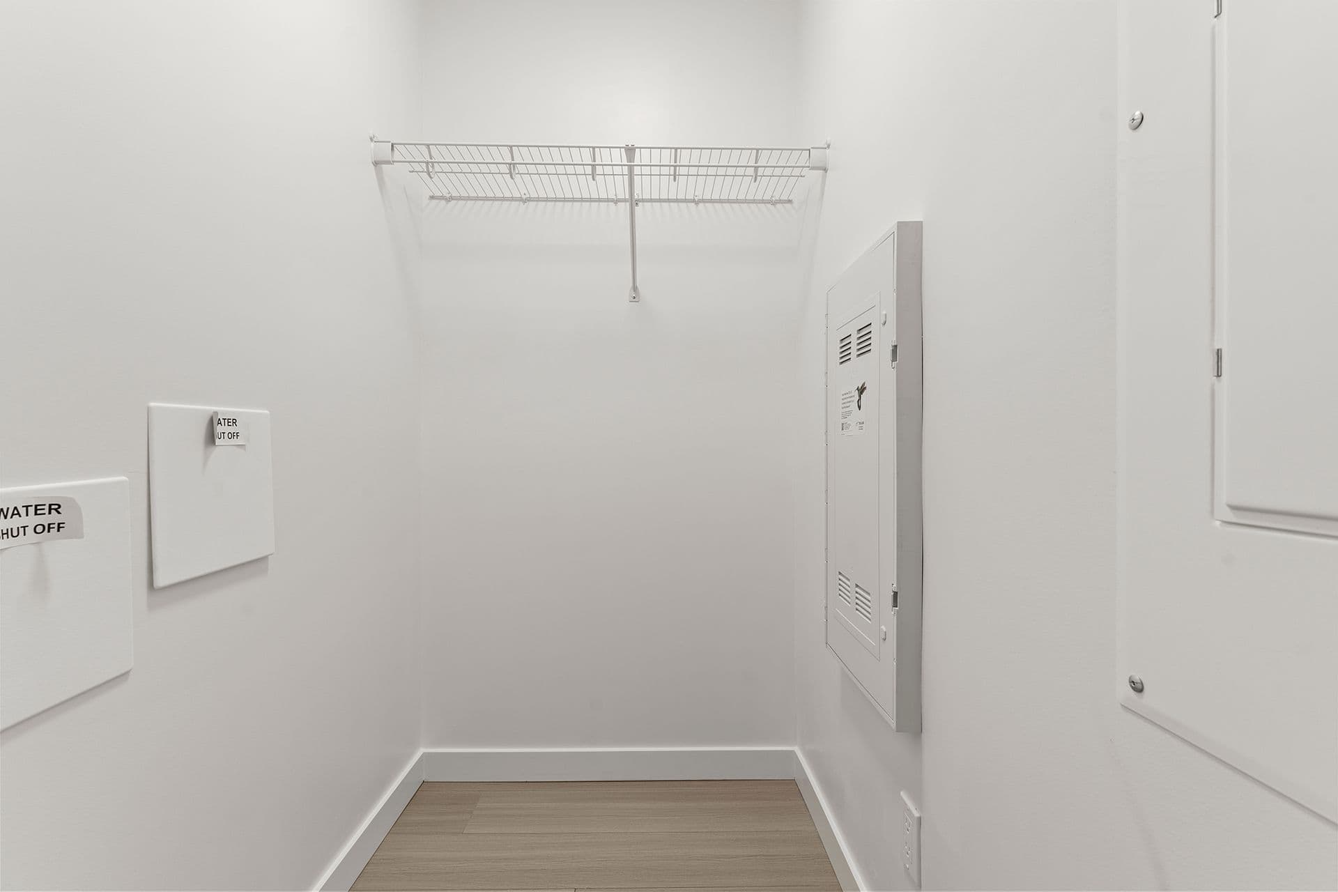 Hawthorne – An empty utility closet with plain white walls, light wood flooring, and a white wire shelf mounted high on the back wall. Access panels, including ones labeled for water shut off, are mounted along the surrounding walls under bright indoor lighting.