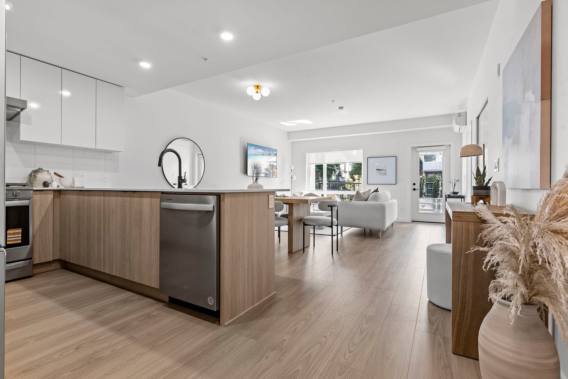 Hawthorne – A modern open-concept kitchen and living space featuring light wood-grain lower cabinets, white upper cabinetry, stainless steel appliances, and light laminate flooring throughout. The area flows seamlessly past a small dining set into a sunlit living room with a white sofa and glass doors leading to an outdoor patio.