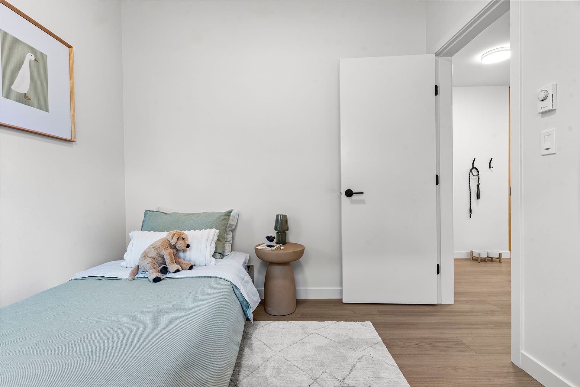 Hawthorne – A bright, minimalist child's bedroom featuring a single bed made with light blue and white bedding, complete with a plush stuffed dog. Modern light wood flooring, plain white walls, a framed duck illustration, and a small round nightstand create a clean, contemporary feel with a view into an adjacent hallway.