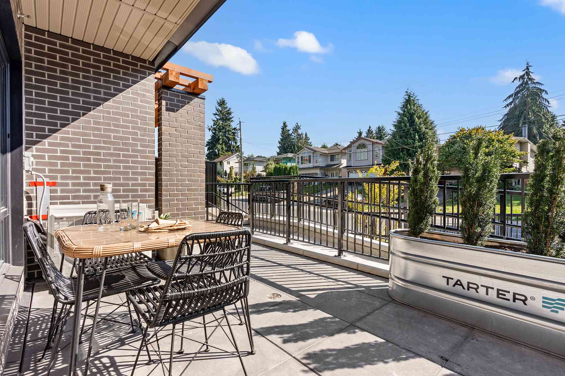 Hawthorne – A sunlit ground-floor patio features a outdoor dining set with woven black chairs, a brick facade wall, and a large metal planter box with small evergreen trees. The patio opens toward a quiet suburban neighborhood street under a bright blue sky.