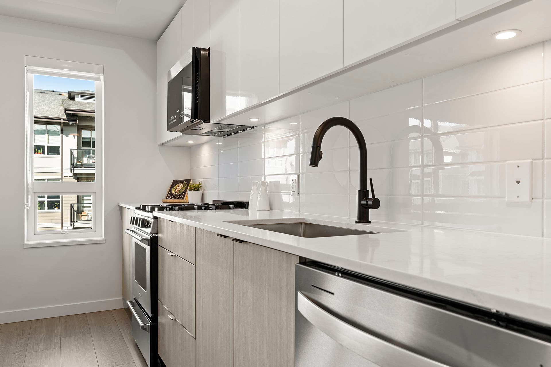 Hawthorne – A modern kitchen featuring light wood lower cabinetry, glossy white upper cabinets, white quartz countertops, and a white subway tile backsplash. Stainless steel appliances, including a gas range stove, microwave, and dishwasher, are paired with a matte black high-arc faucet.