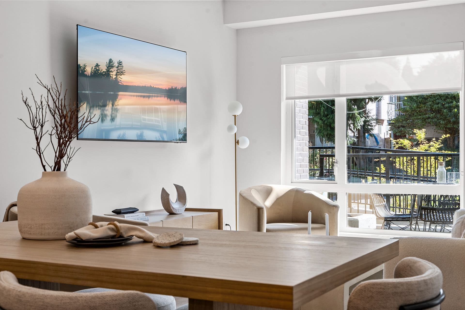Hawthorne – A modern living and dining area featuring a light-wood dining table in the foreground and a wall-mounted television displaying a nature scene. A large window brings in bright daytime light and offers a view of an outdoor patio area with green trees.