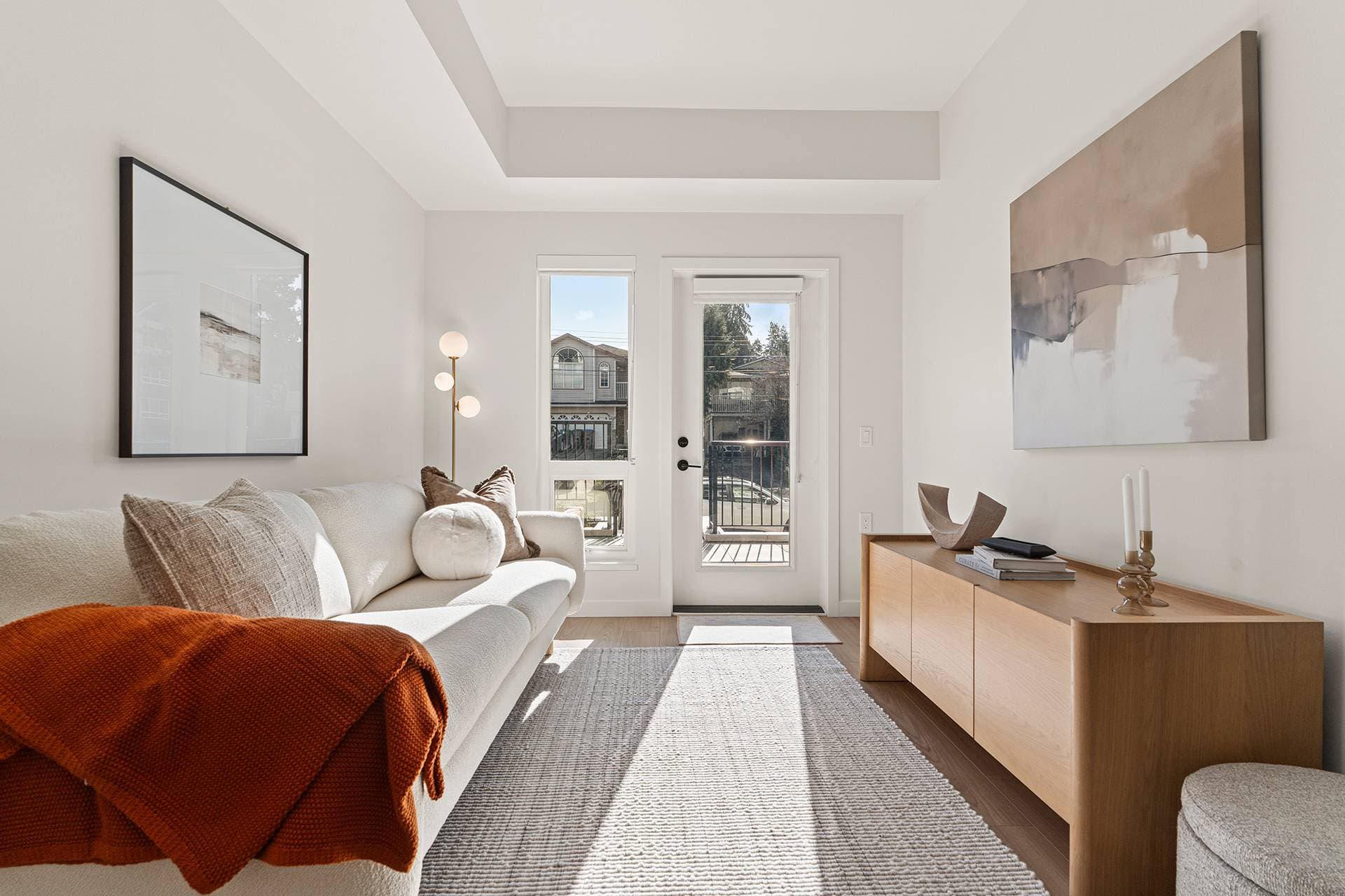 Hawthorne – A bright, modern living room features a cream-colored sofa draped with a rust-orange throw blanket, a textured area rug over light wood flooring, and a low wooden sideboard against white walls. Large glass doors and tall windows let in abundant daylight while offering a view of surrounding residential buildings outside.