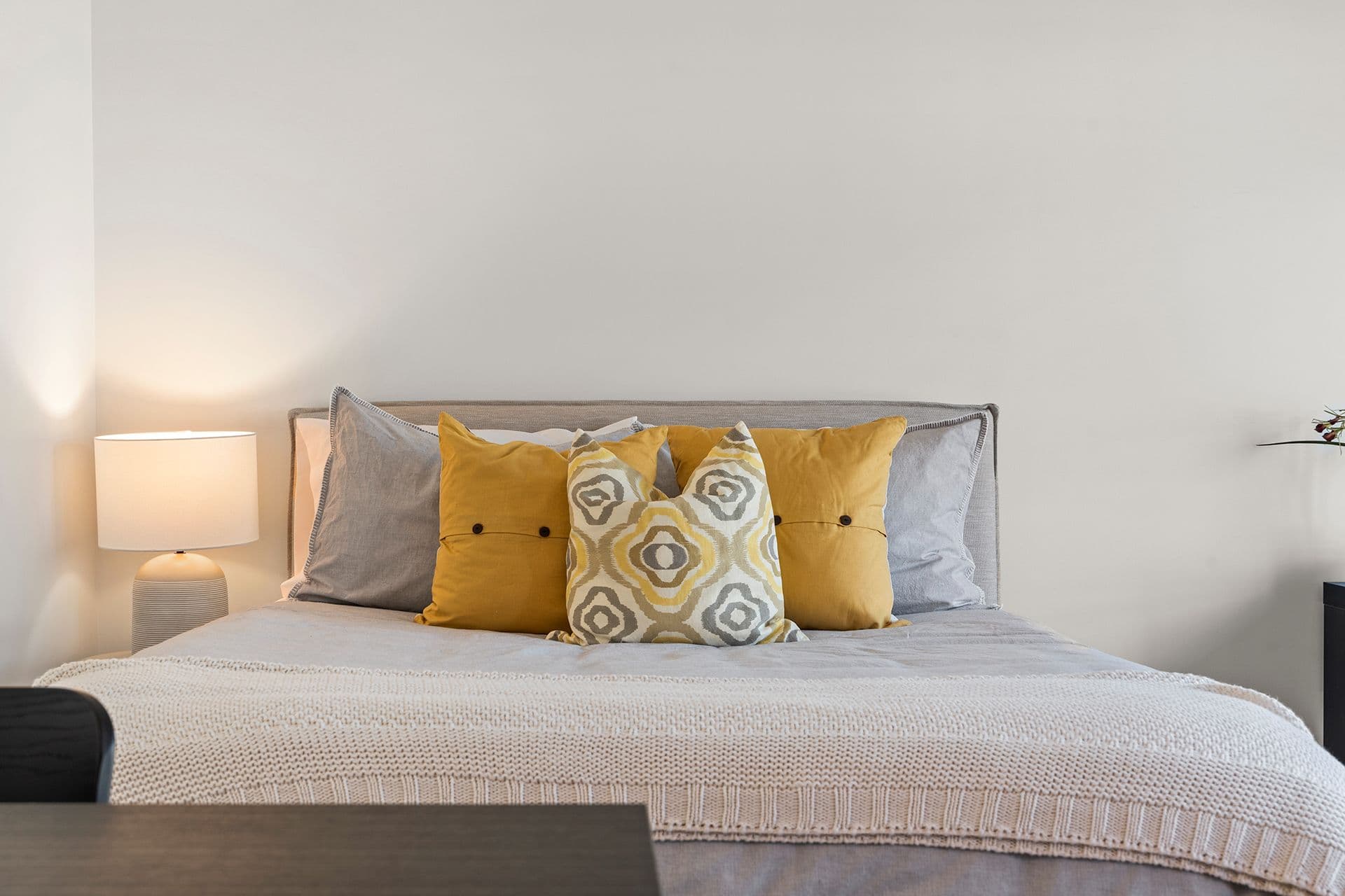 Hawthorne – A neatly dressed bed with a light grey upholstered headboard features grey linens, vibrant mustard-yellow accent pillows, and a geometric pattern cushion. Beside the bed, an illuminated table lamp with a ceramic base sits against a plain white wall, creating a warm, cozy atmosphere.