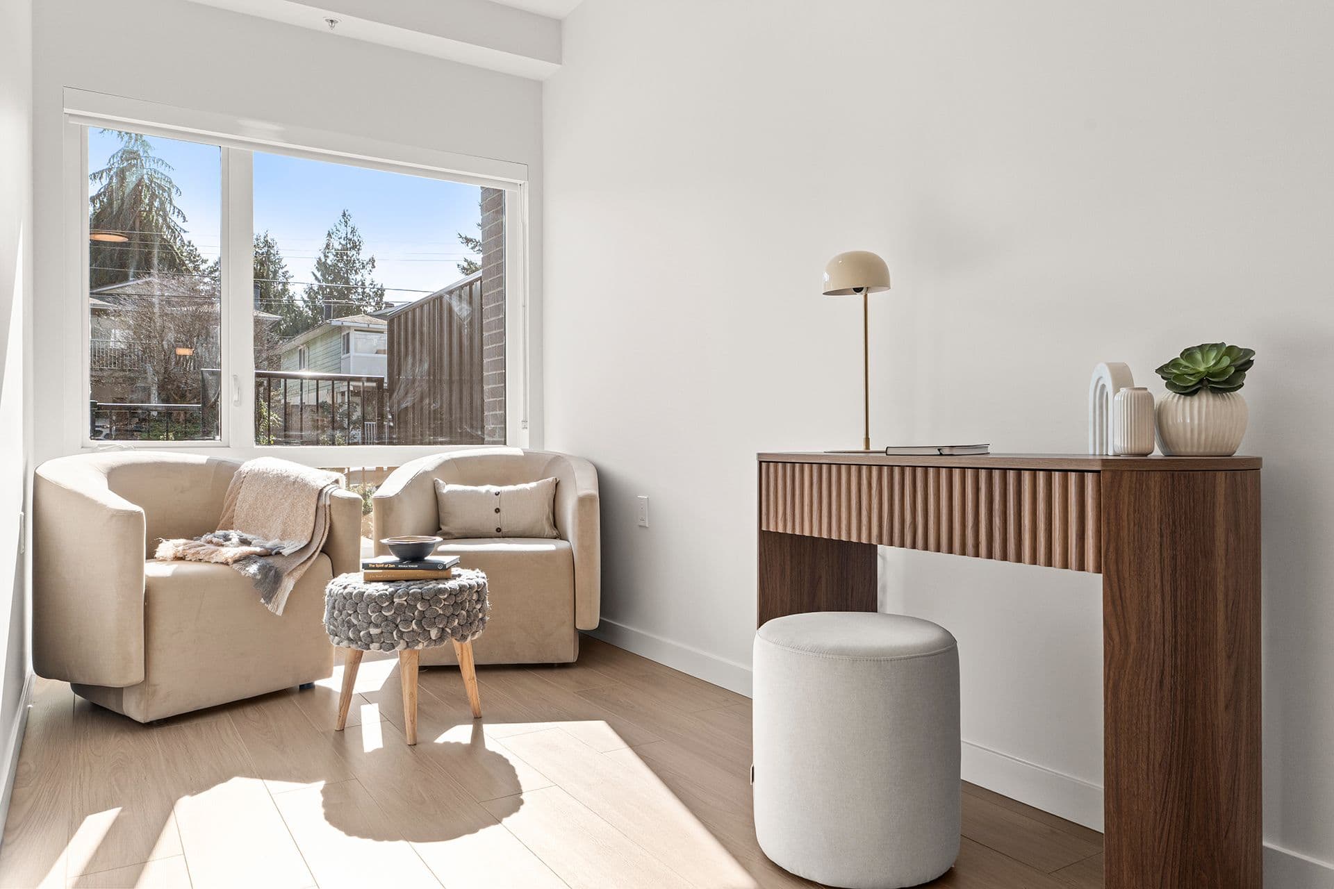 Hawthorne – A sunlit interior room featuring light hardwood flooring, white walls, and a cozy seating nook with two beige barrel armchairs positioned by a large window. Adjacent to the seating area is a fluted wooden console desk equipped with a round stool, minimalist table lamp, and decorative accent pieces.