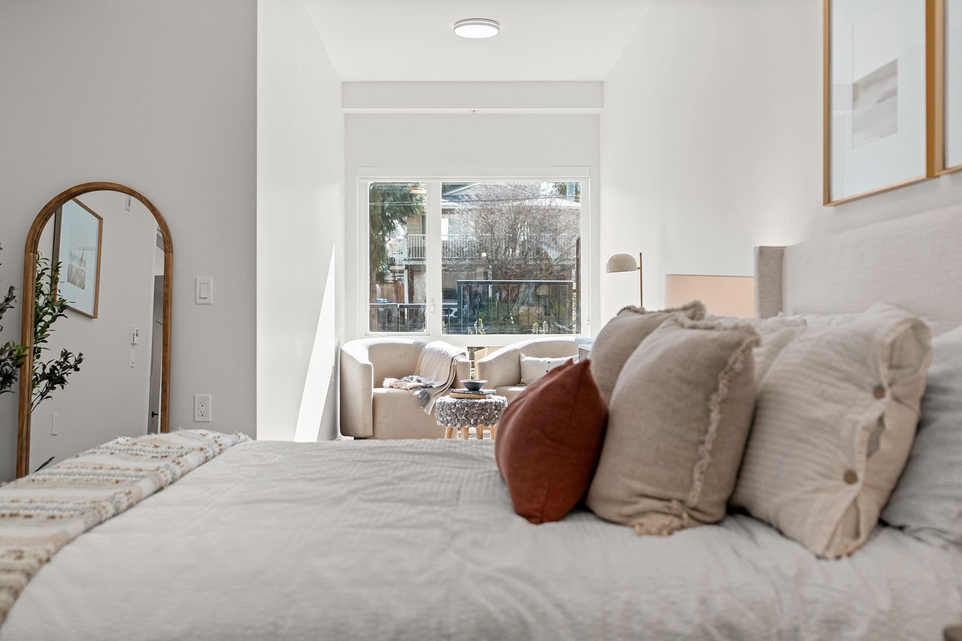 Hawthorne – A bright, modern bedroom featuring a bed dressed in neutral linens and decorative pillows in the foreground, with an arched full-length floor mirror along the left wall. In the alcove near the window, two plush armchair seats framing a small side table create a cozy reading nook filled with natural daylight.