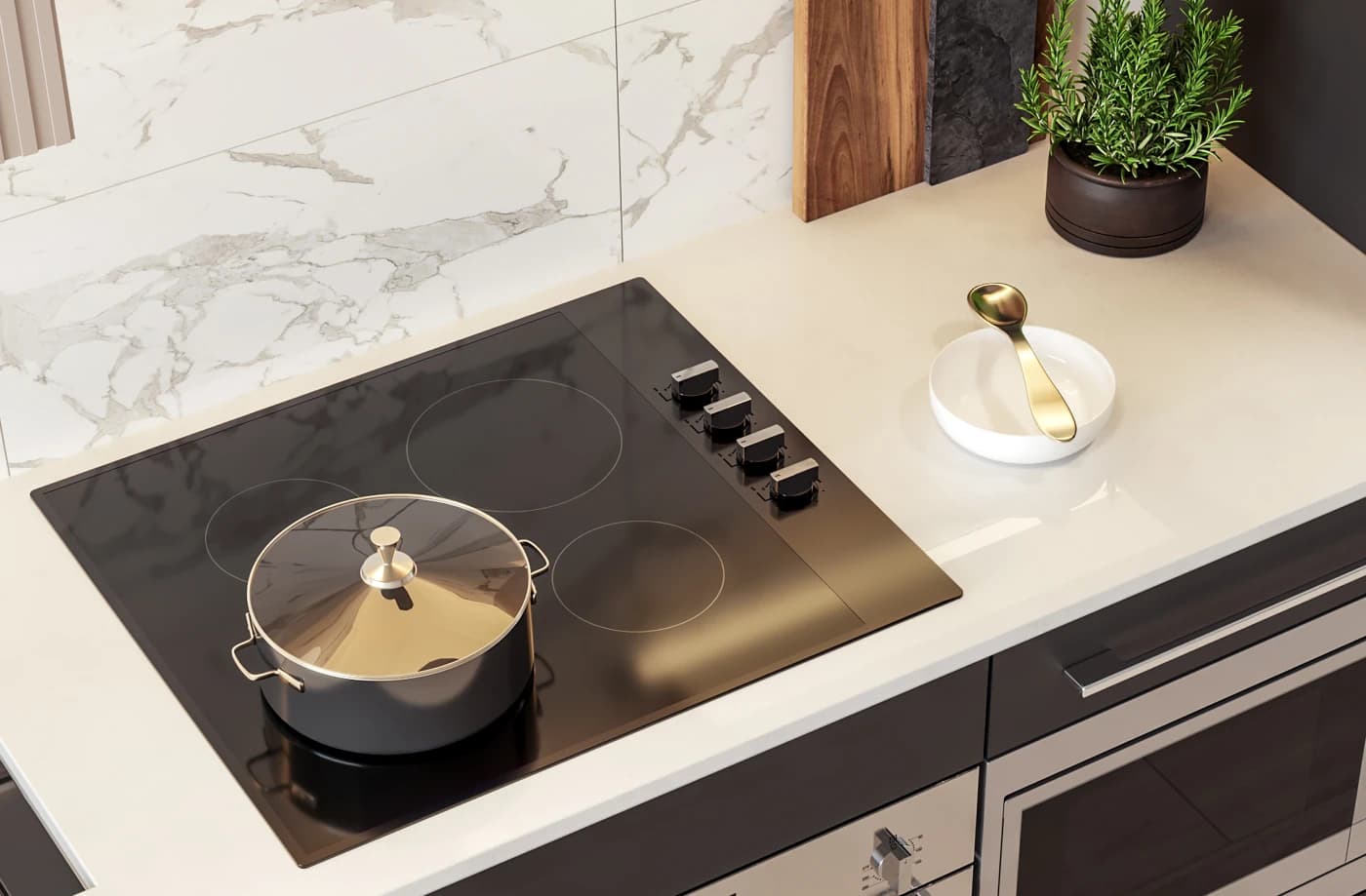 Piano – A high-angle close-up of a sleek black glass electric cooktop set into a white quartz countertop with dark cabinetry beneath. The kitchen scene features a stainless steel pot, a small potted plant, a white marble backsplash, and a gold serving spoon resting nearby.