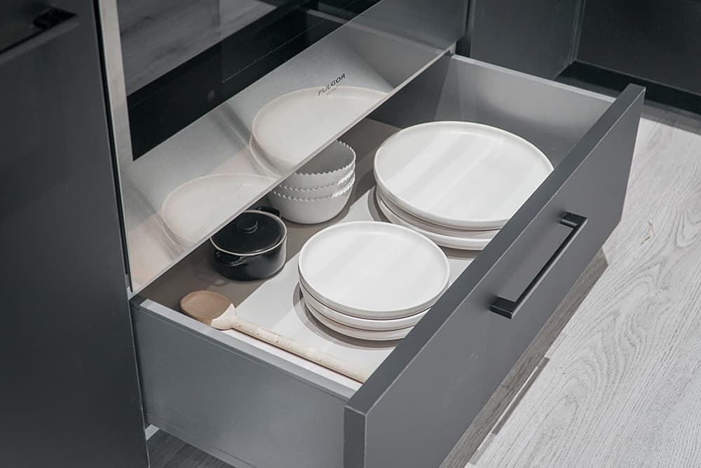 Piano – A detailed view of an open charcoal-grey kitchen drawer positioned beneath a stainless steel Fulgor Milano oven. The drawer is neatly organized with stacks of modern white ceramic plates, bowls, a small black pot, and a wooden spoon against light wood-look flooring.