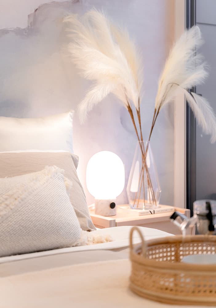 Piano – A cozy bedroom vignette features neutral-toned pillows and a woven tray in the foreground. In the background, a light wood nightstand holds a glowing orb lamp and a glass vase with fluffy pampas grass against a soft watercolor accent wall.