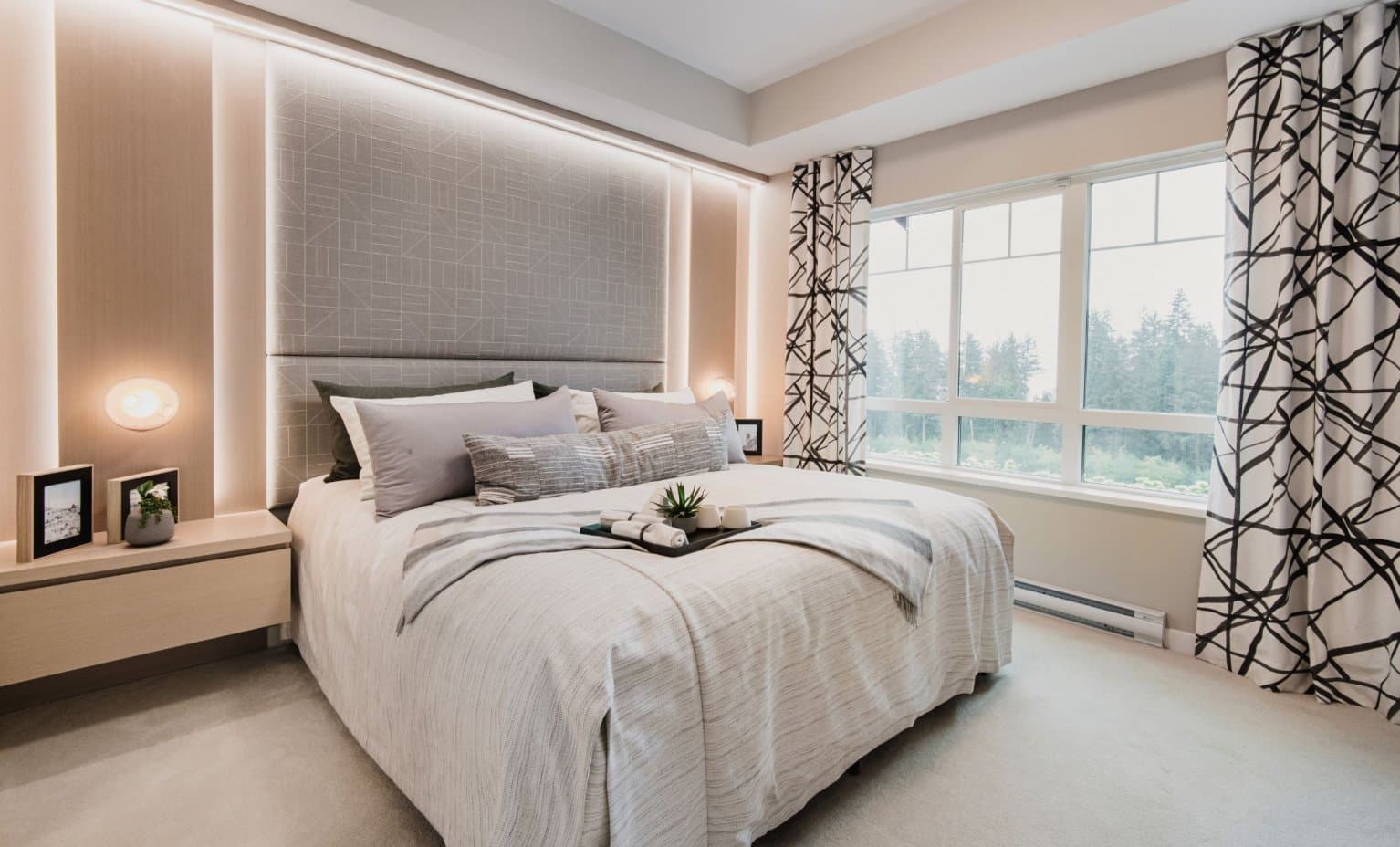 Partington Creek – Bedroom with a padded grey upholstered headboard wall, a made bed with a service tray, and large windows overlooking trees.