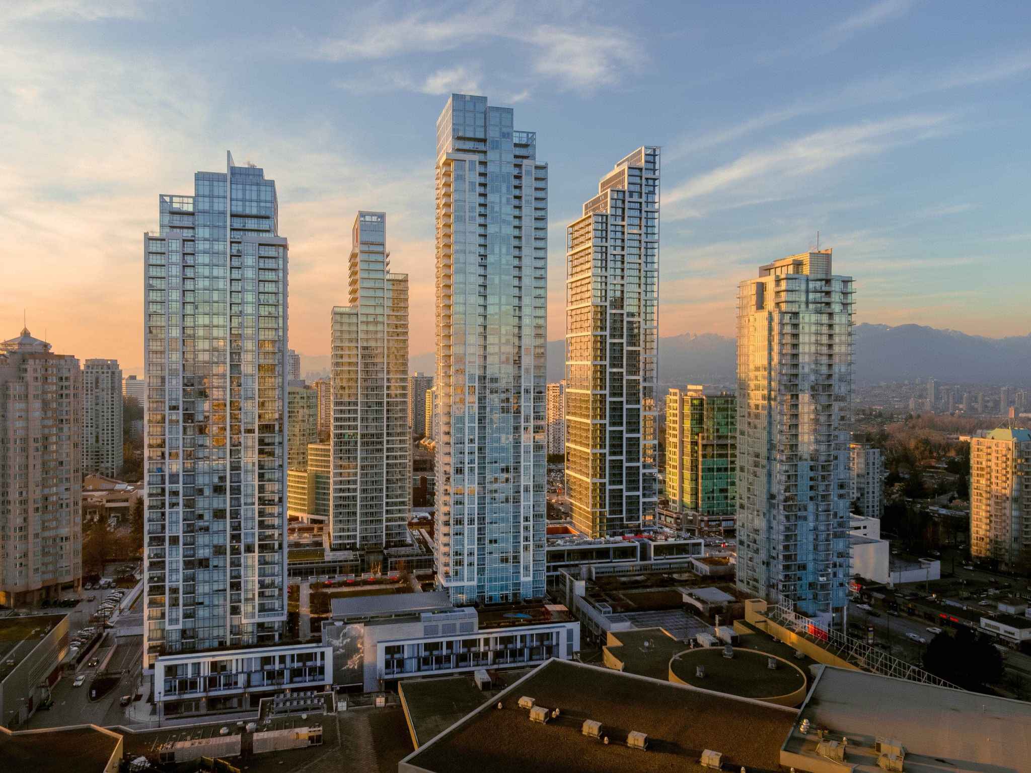 Ethos – An elevated drone perspective showcases a cluster of modern glass high-rise residential towers rising above a multi-level podium with green rooftops. Warm golden sunset light illuminates the building facades, casting long reflections with distant mountain silhouettes and a soft sky visible in the background.