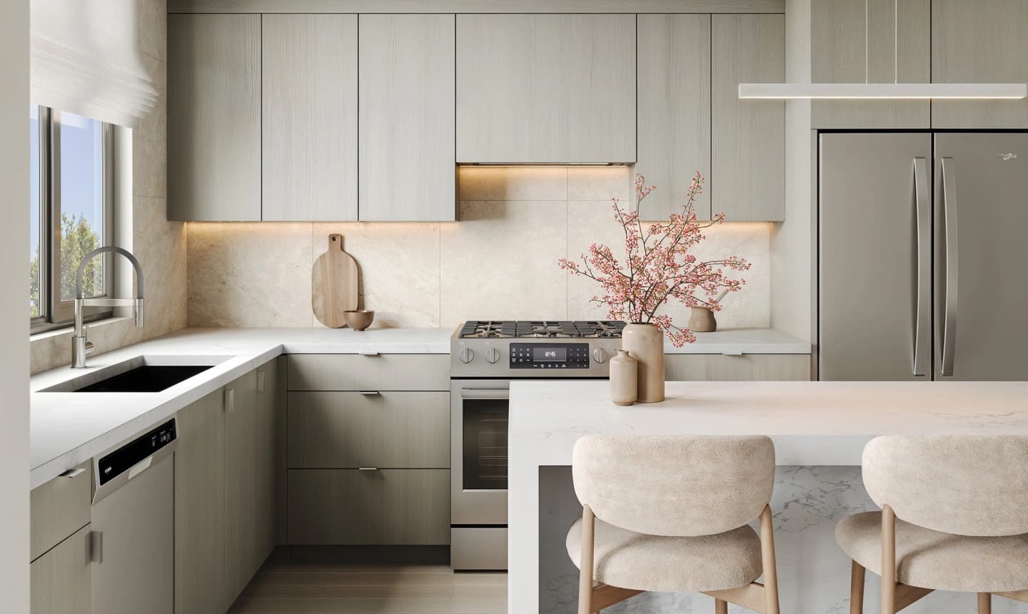 Camden by the Parks – A modern, bright kitchen featuring light wood-grain flat-panel cabinetry with soft under-cabinet lighting, stainless steel appliances, and white marble countertops. A waterfall kitchen island is paired with upholstered bar stools and decorated with a vase of pink blossom branches.