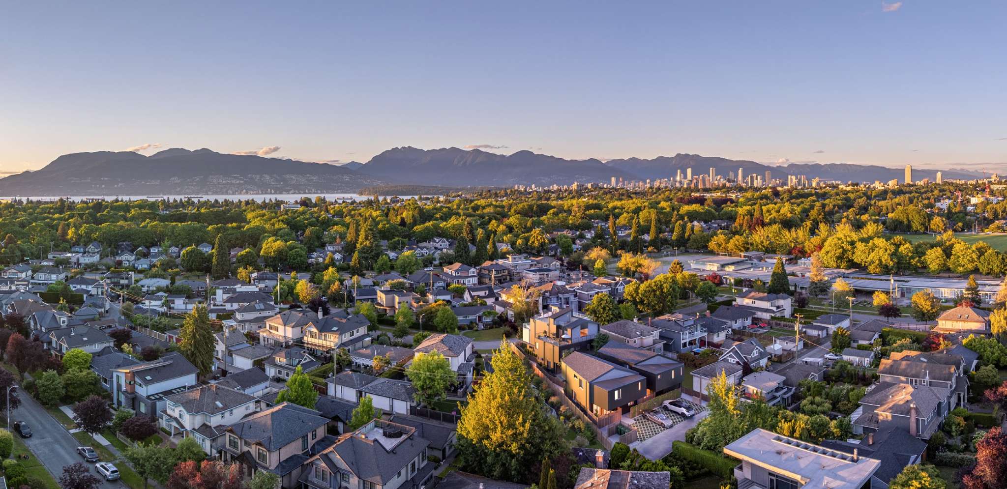 Katsura – A high-angle aerial view looks across a leafy residential neighbourhood of single-family homes bathed in warm golden-hour sunlight. In the distance, a calm body of water leads to a distant city skyline framed by a sprawling mountain range under a clear sky.