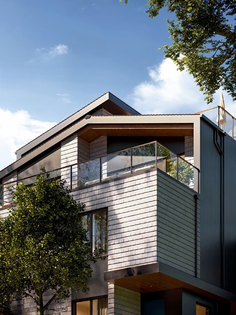 Katsura – A detailed architectural view of the upper levels and pitched roof of a modern residential building, featuring light-toned brick or tile cladding, dark vertical siding, and a private balcony with glass railings. Lush green tree branches frame the structure against a bright, clear blue sky with soft white clouds.