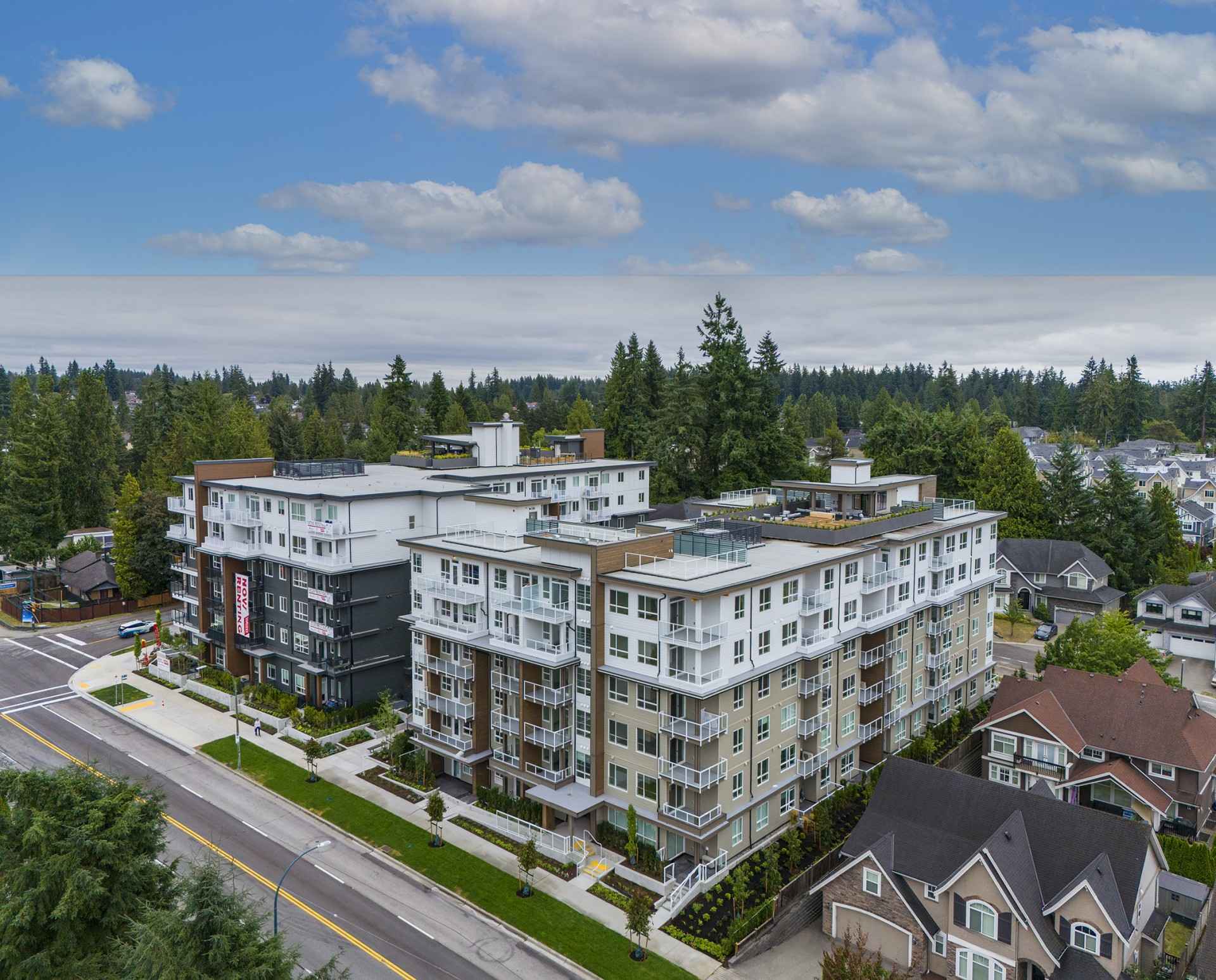 Sol – An elevated drone shot shows a modern, multi-story residential building complex with white and beige paneling, private glass balconies, and rooftop patios. The surrounding suburban neighborhood features paved streets, detached single-family homes, and a dense forest of evergreen trees under a blue sky with scattered clouds.