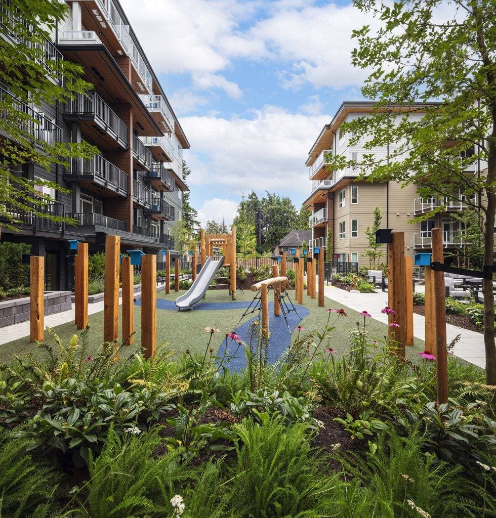 Sol – A outdoor children's playground equipped with wooden posts, a metal slide, and climbing structures, situated on a soft green and blue safety surface between modern residential buildings. Lush green gardens with flowering plants frame the foreground under a bright, partly cloudy daytime sky.