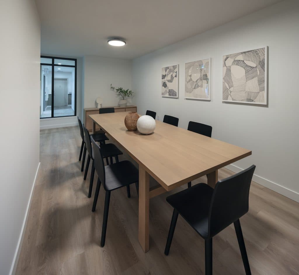 Sol – A modern communal dining room or boardroom featuring a long light-wood table paired with sleek black chairs under soft indoor lighting. Light oak flooring, neutral white walls decorated with framed abstract artwork, and a glass partition looking into a hallway create a clean and sophisticated atmosphere.