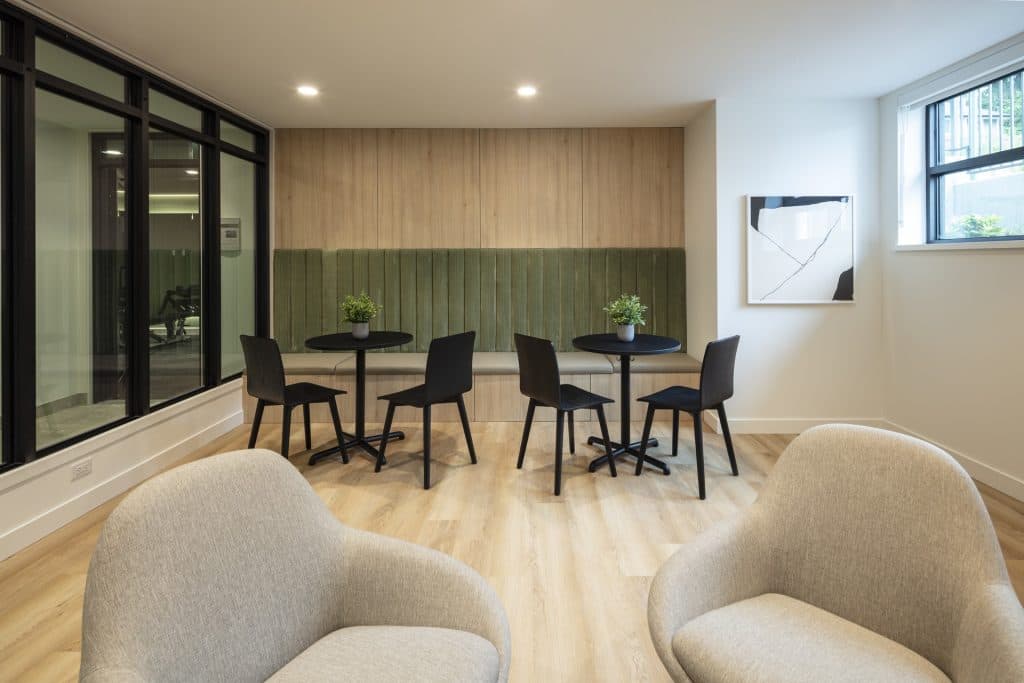 Sol – A modern indoor lounge area features a built-in wooden banquette with sage green upholstered padding, paired with small black bistro tables and matching chairs. Light wood flooring, glass-partitioned adjacent rooms, and plush grey armchairs in the foreground create a warm, inviting communal space.