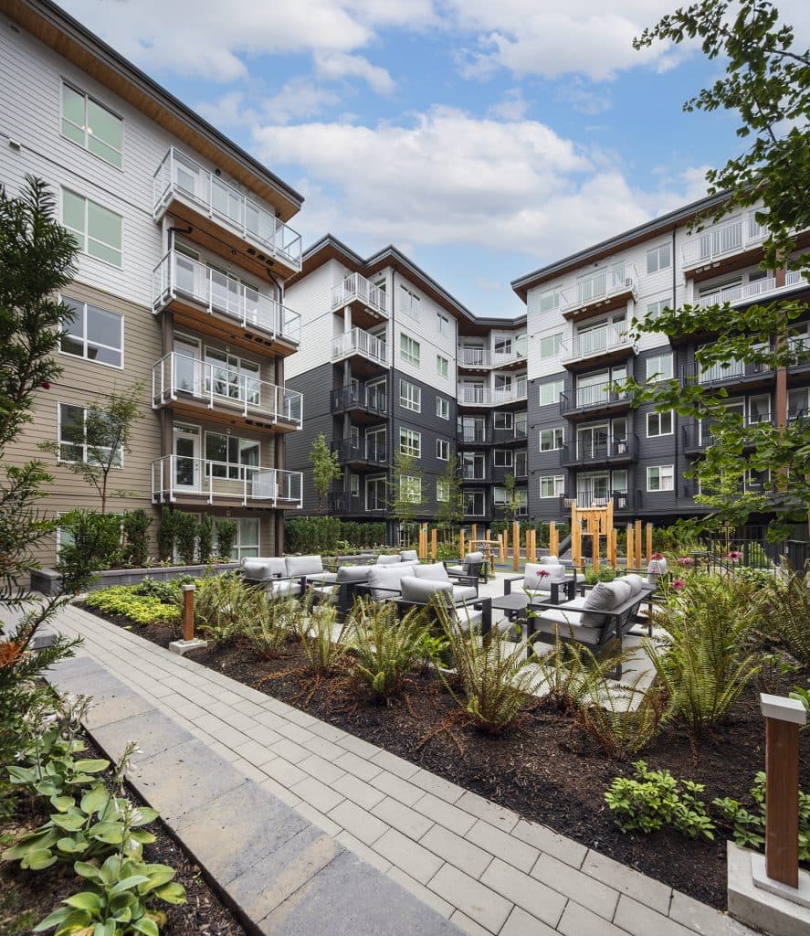 Sol – A ground-level outdoor courtyard features cushioned lounge seating and a paved walkway surrounded by lush landscaping, ferns, and garden beds. In the background, multi-story residential buildings with private balconies encircle the courtyard and a wooden children's play structure under a bright, partly cloudy sky.