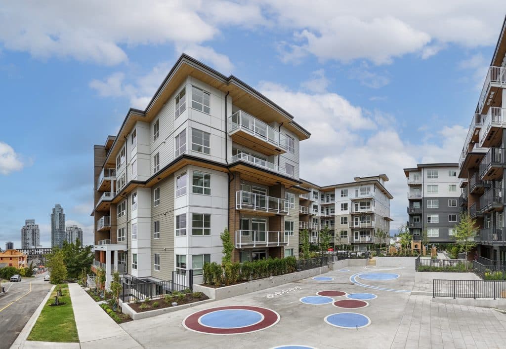 Sol – A modern low-rise condominium building featuring light siding, wood-toned soffits, and private balconies overlooks a concrete courtyard with painted circular designs. The scene is shot on a clear day with a bright blue sky, revealing a residential street slope and high-rise towers in the distant background.