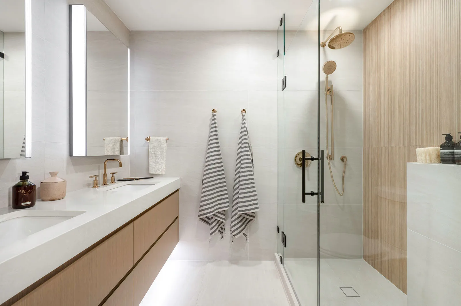 Rhodes – Spa-style bathroom with double vanity, backlit mirrors, brass fixtures and a glass walk-in shower with wood-look tile feature wall.