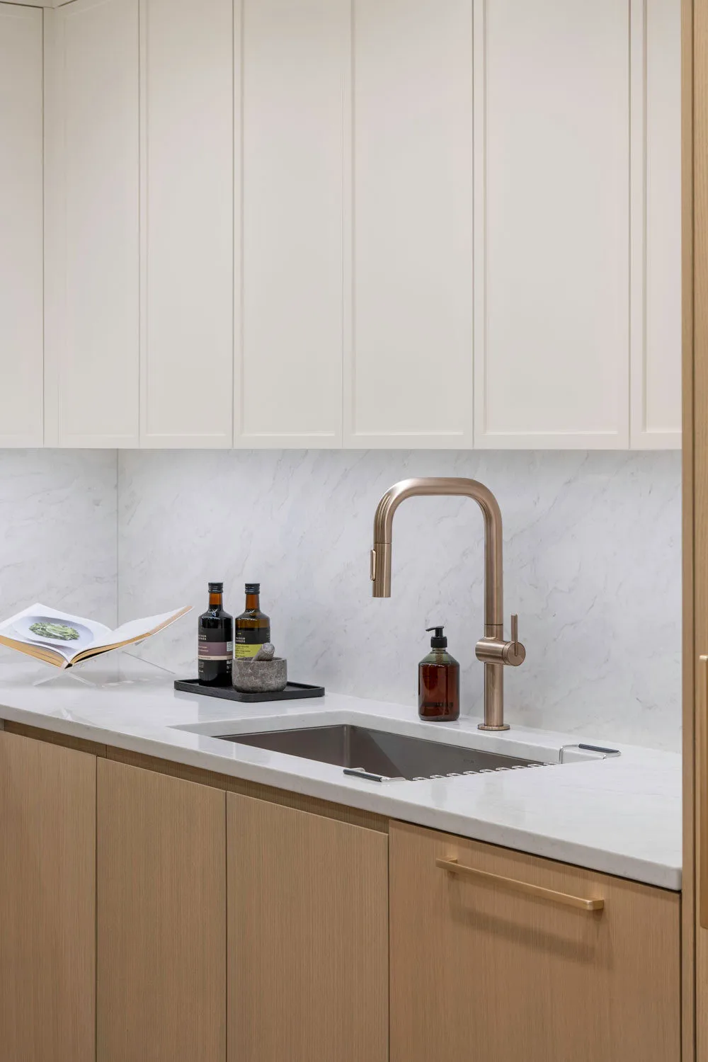 Rhodes – Kitchen sink detail with brushed-brass pull-down faucet, marble backsplash and cream shaker upper cabinets.