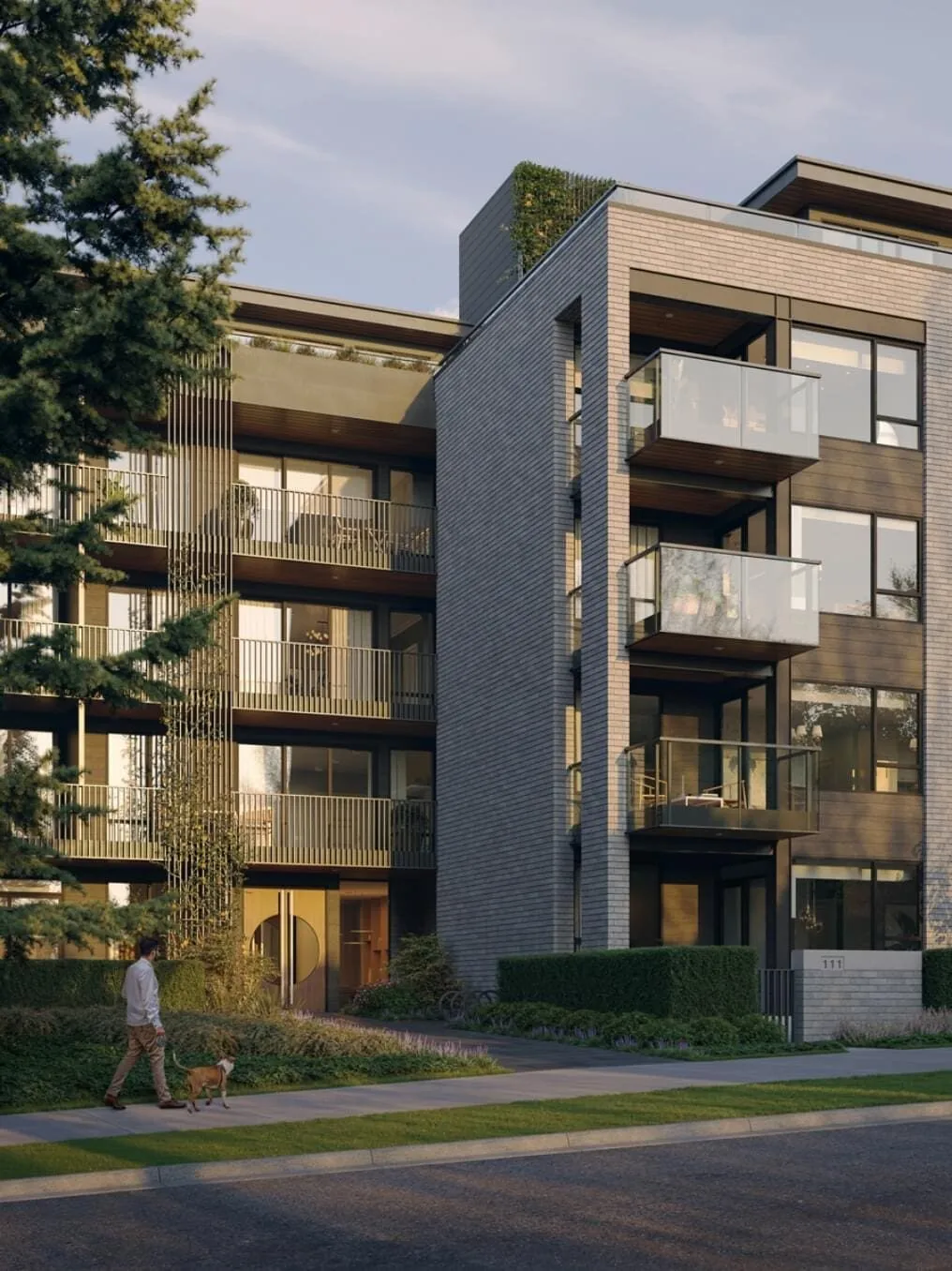Rhodes – Portrait street-level view of the brick facade at dusk with glass balconies, warm lobby entry, landscaped frontage and a man walking a dog on the sidewalk.