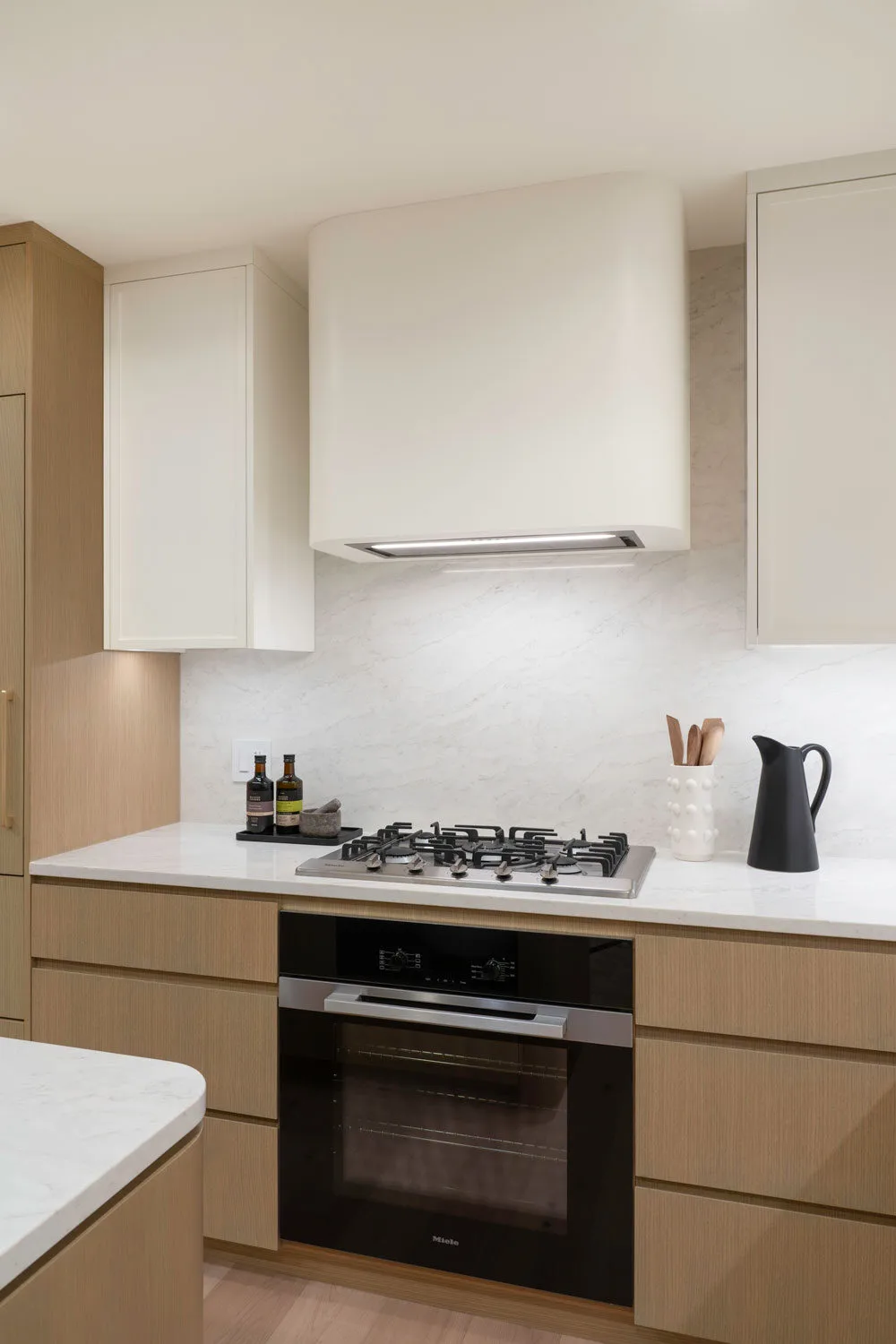 Rhodes – Kitchen detail of the gas cooktop and built-in Miele wall oven set in light-oak cabinetry with marble backsplash.
