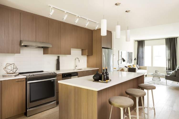 Allwood Place – Kitchen with dark wood cabinetry, stainless appliances, track lighting, and an island with wood stools opening onto a living room.