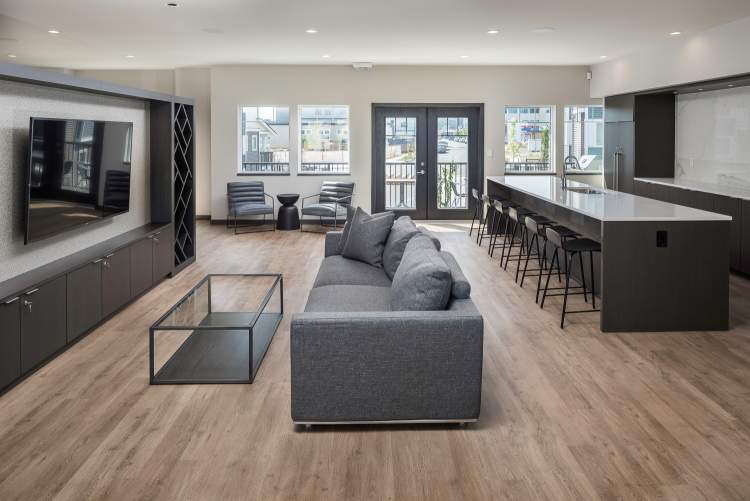 Allwood Place – Shared lounge/clubroom with a wall-mounted TV and dark built-in shelving, a grey sofa, and a kitchenette island with bar stools opening to a balcony.