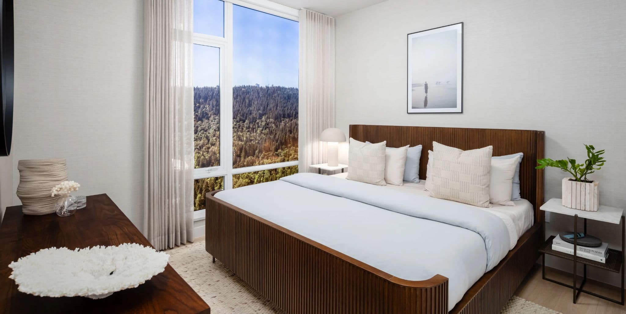Ironwood – A bedroom with a dark wood ribbed bed frame, layered white and blue pillows, and a large window looking out over treetops.