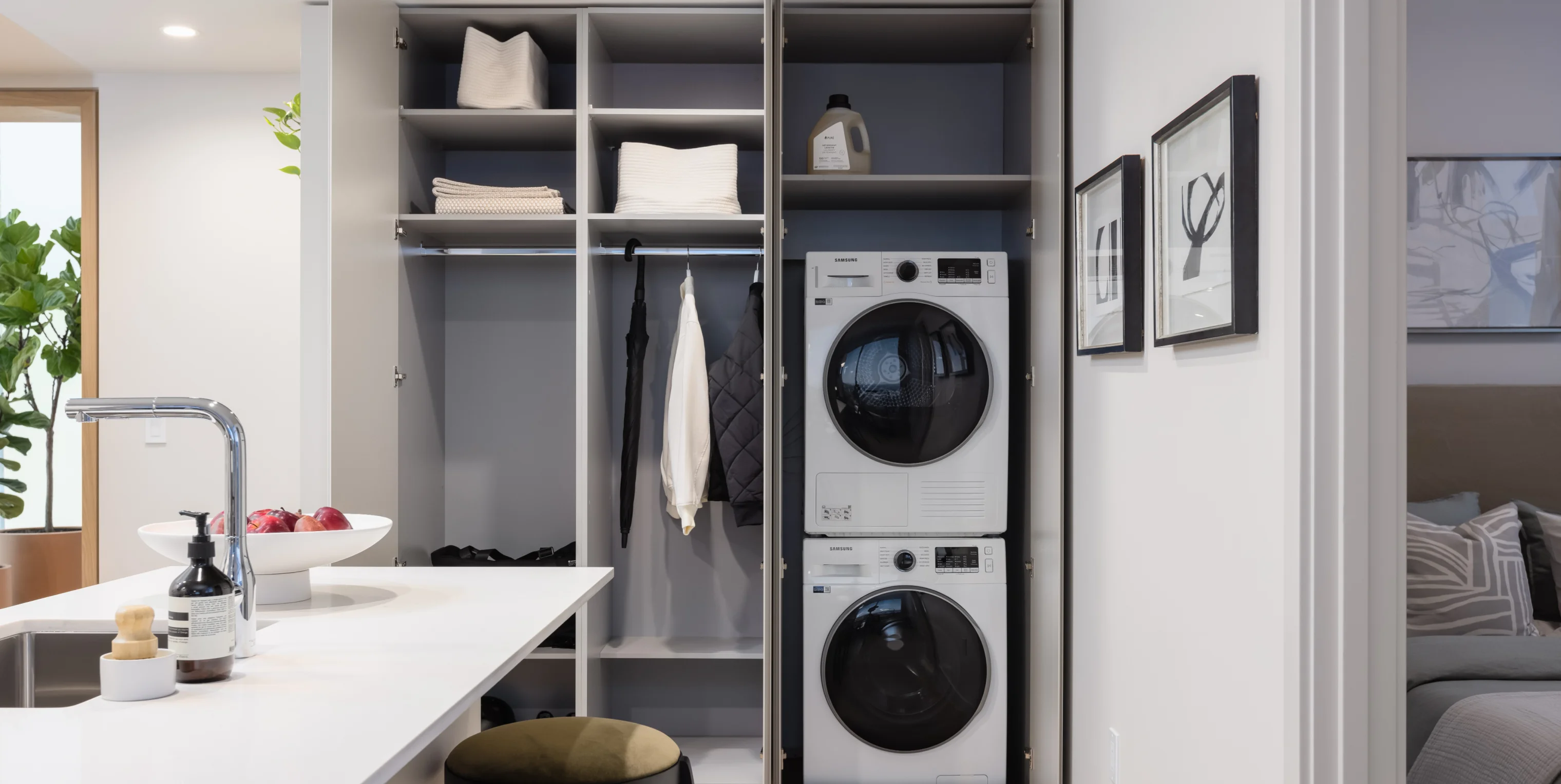 Ironwood – An open closet and mudroom area with stacked washer and dryer, shelving with linens, hanging coats, and a kitchen counter with a fruit bowl in the foreground.
