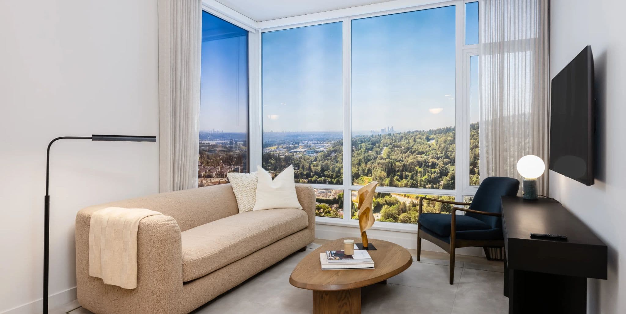 Ironwood – A corner living room with a beige sofa, navy accent chair, wood coffee table, and floor-to-ceiling windows overlooking a forested valley and distant skyline.