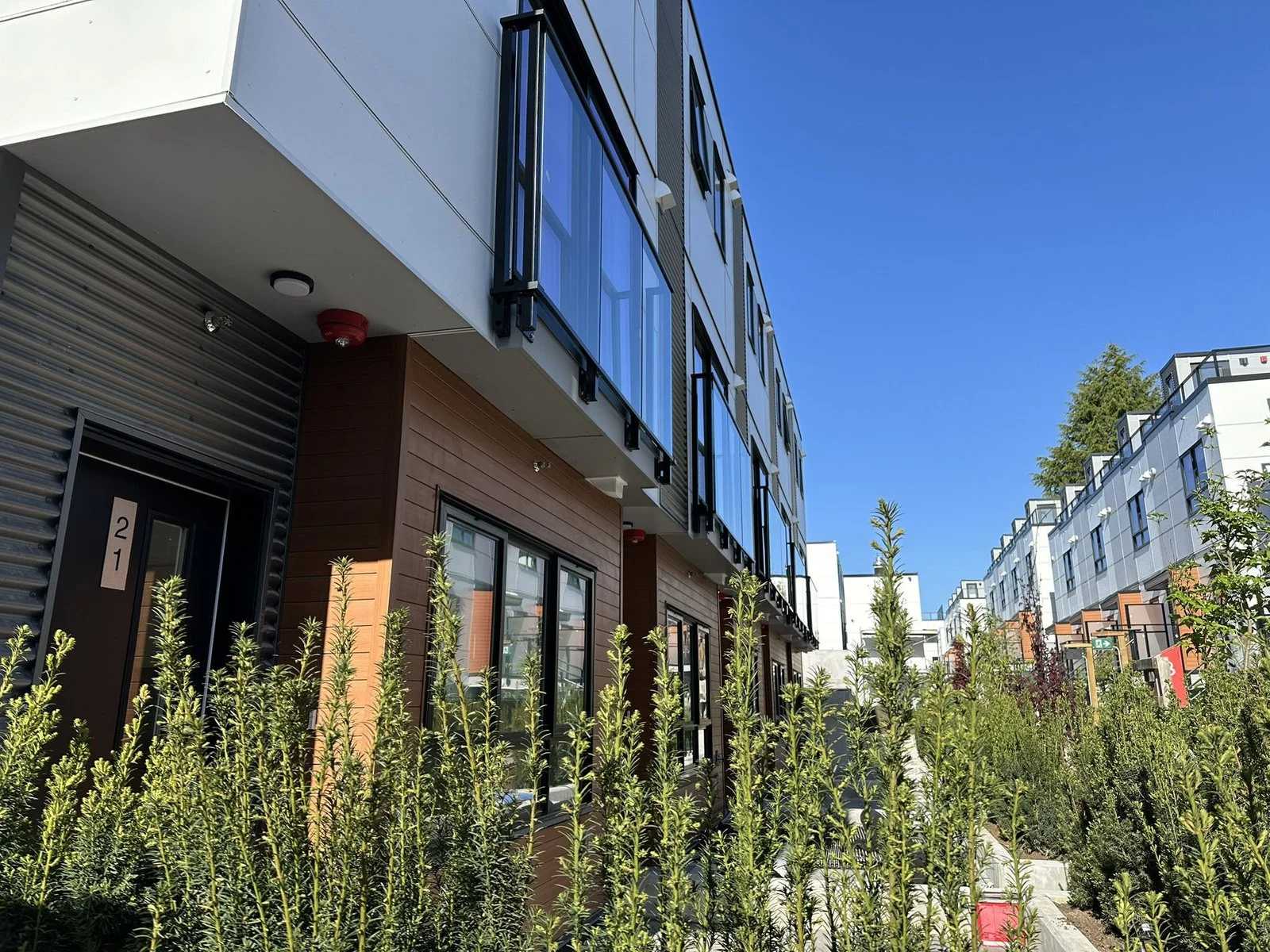 Lilibet – Close-up exterior view of a townhome unit entrance (No. 21) with wood-look siding, corrugated metal accent, glass balcony railings, and landscaping of young shrubs.