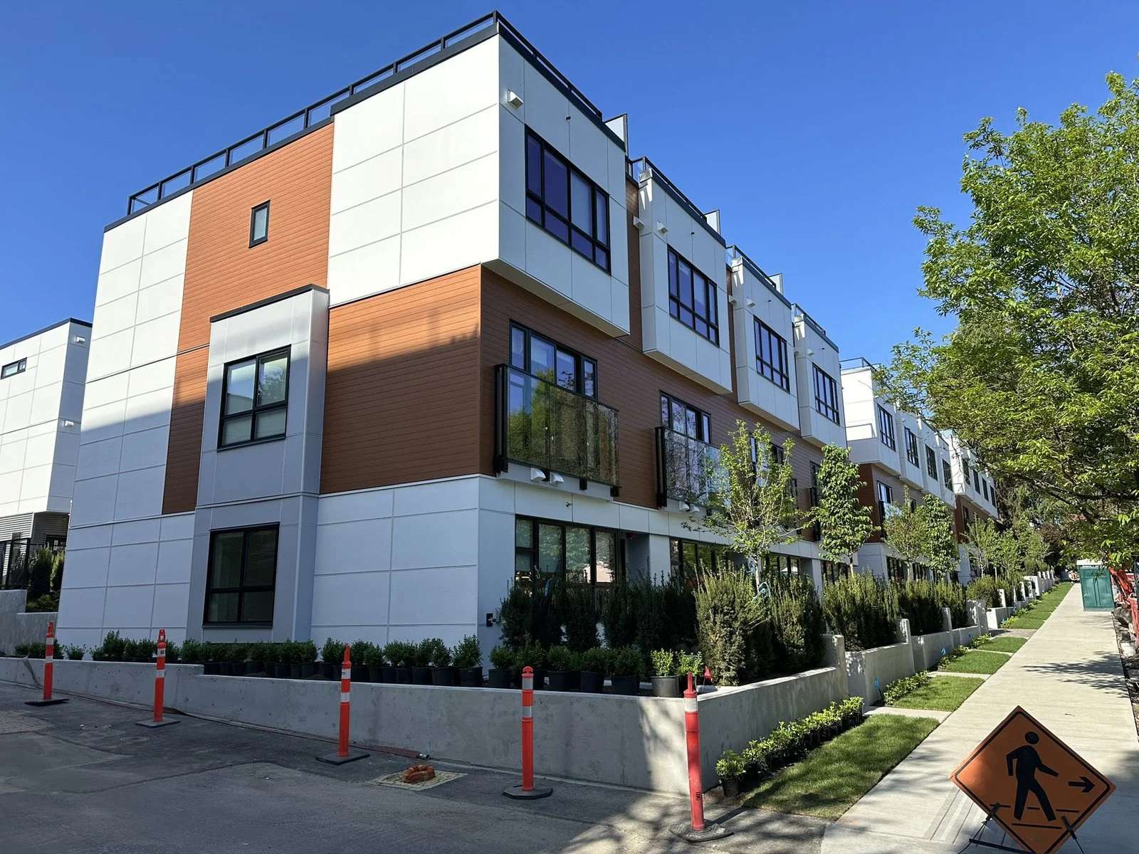 Lilibet – Corner exterior view of a townhome building with white, grey, and burnt-orange wood-look siding, black-framed windows, and a landscaped perimeter with orange traffic delineators from ongoing construction.