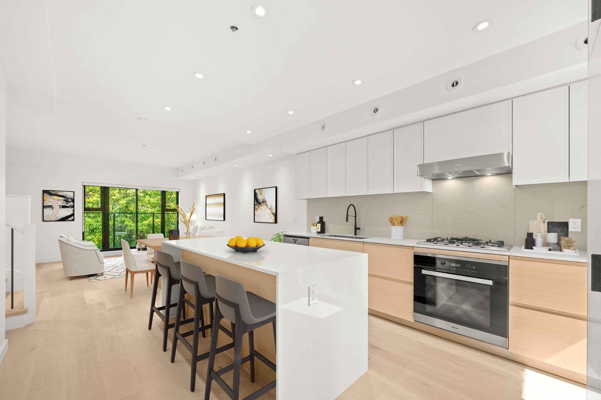 Lilibet – Kitchen with white and light wood cabinetry, a waterfall island with bar seating, stainless gas cooktop and built-in oven, opening to a dining and living area.