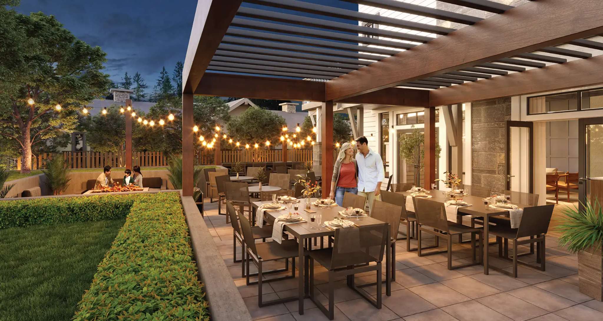 Terrayne Towns – An outdoor patio and dining terrace under a dark wooden pergola is filled with long tables, setting places, and warm string lights at dusk. People are gathered, including a couple standing together and a group seated near a fire pit in the background amidst manicured lawns and trees.