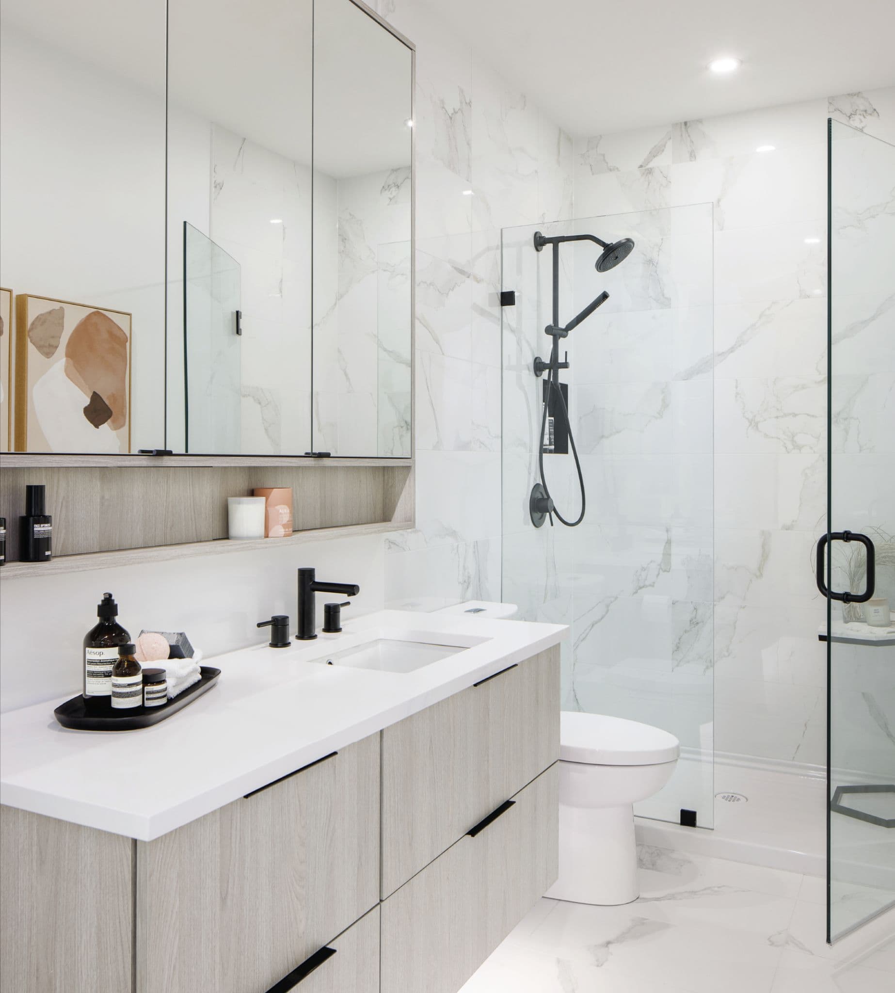 Terrayne Towns – A modern bathroom featuring a floating light wood vanity with a white countertop, matte black hardware, and mirrored medicine cabinets above. White marble-style porcelain tiles line the walls and floor, extending into a frameless glass-enclosed shower with black fixtures.