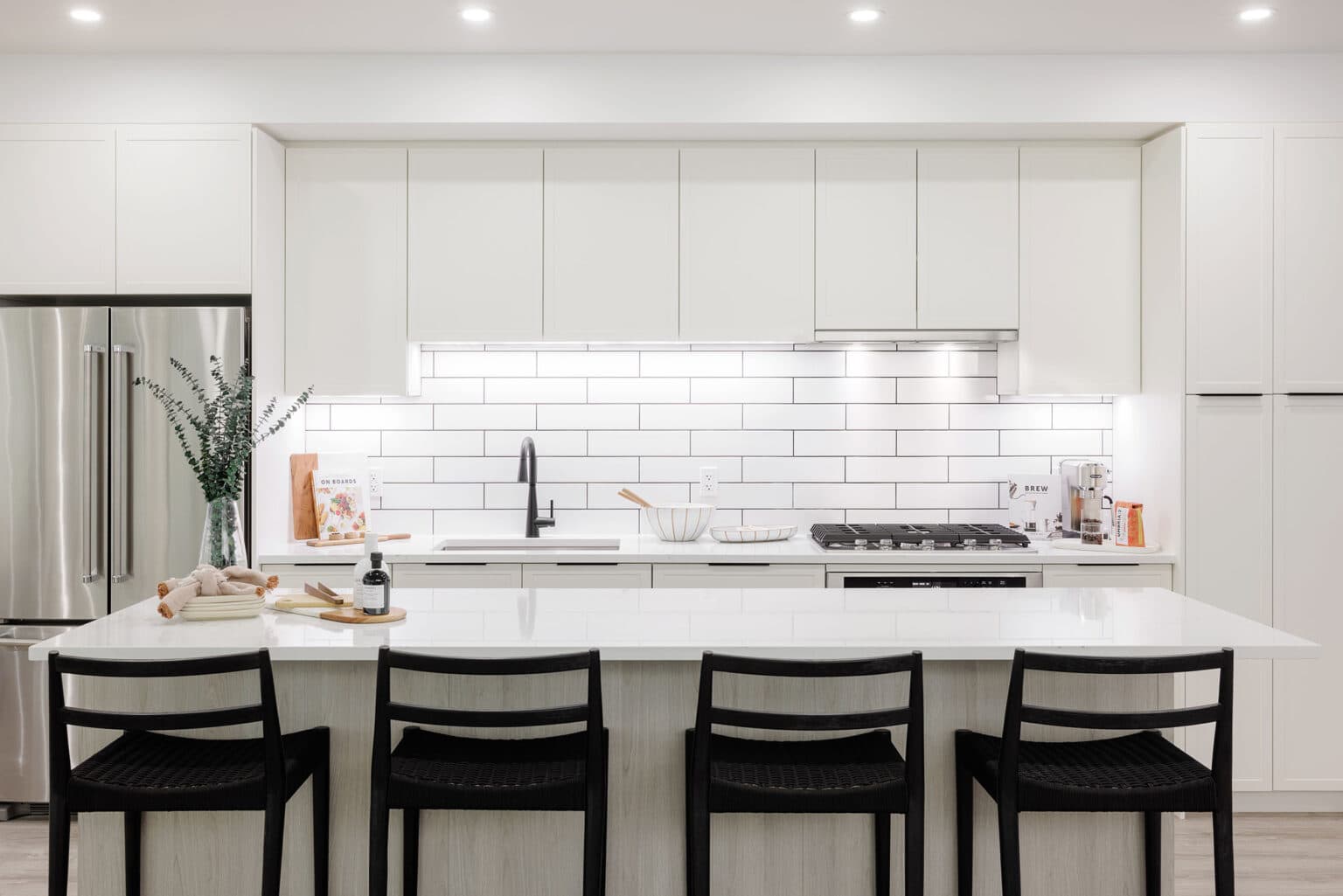 Terrayne Towns – A modern, bright kitchen featuring seamless white flat-panel cabinetry, a white subway tile backsplash with under-cabinet lighting, and stainless steel appliances. A large central island with a solid white countertop and light wood base is lined with four black woven-seat barstools and styled with subtle decor objects.