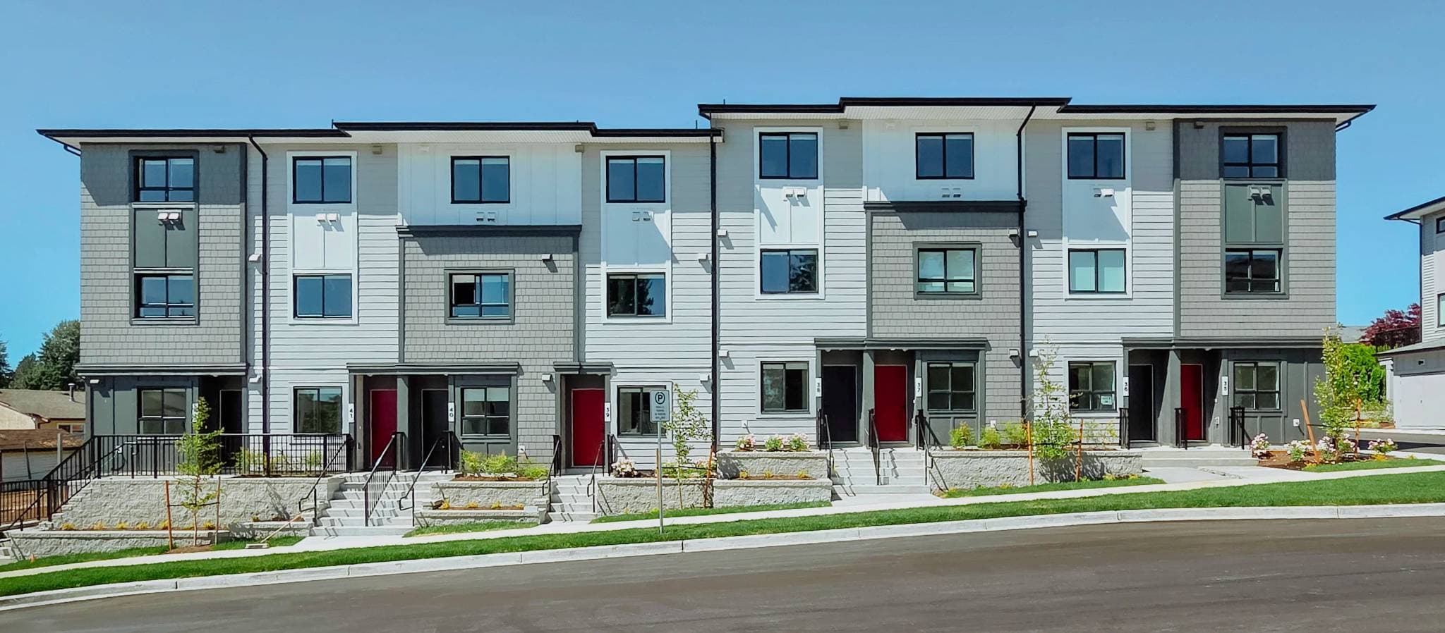 South Grove – A row of modern three-story townhomes features alternating white and gray horizontal siding with shake-shingle accents and bold red front doors. Elevated entrances with short concrete stairs and retaining walls face a freshly paved street beneath a clear blue sky.
